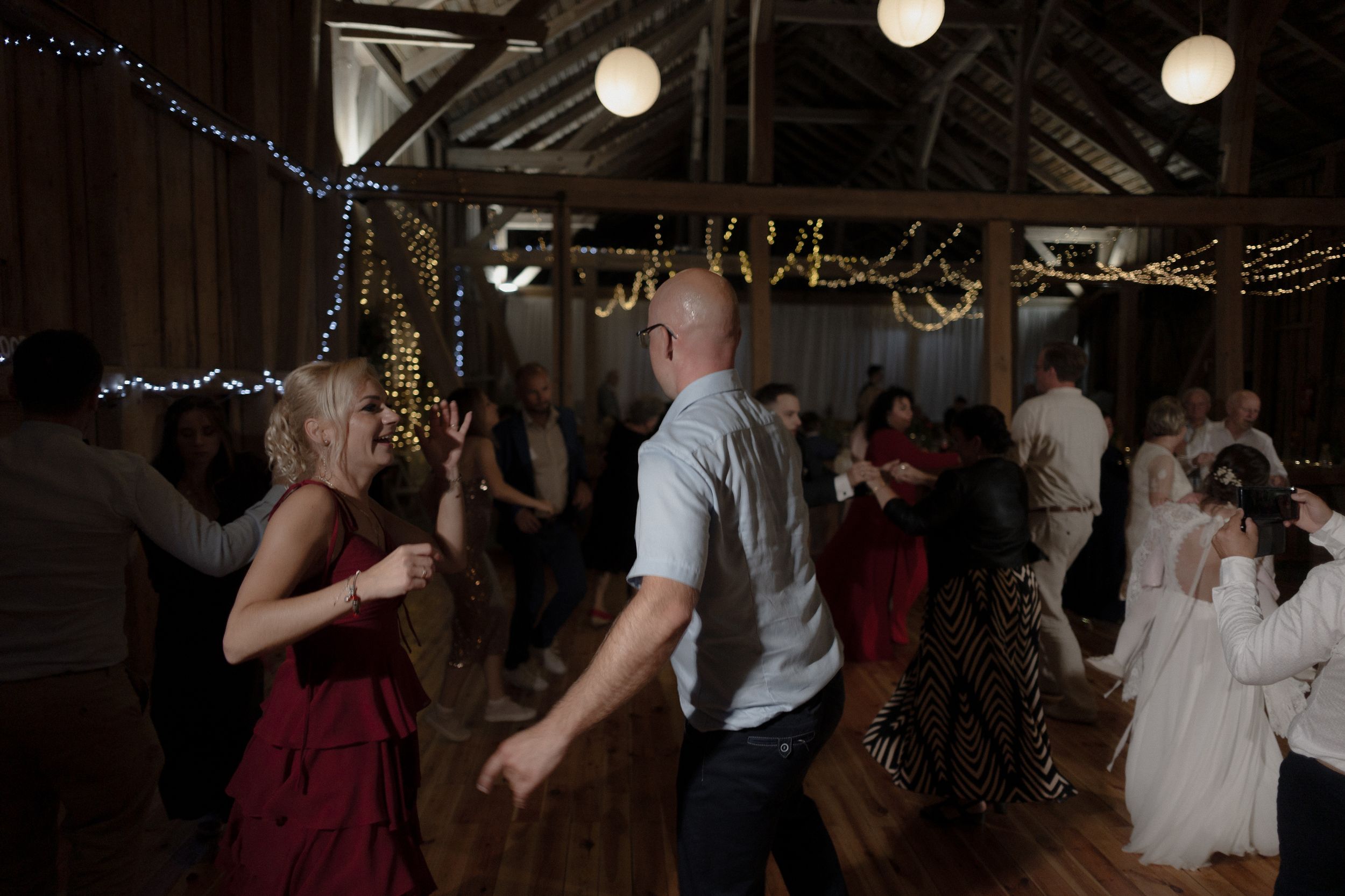 Guests dancing and celebrating at a dimly lit indoor wedding reception venue with string lights overhead.
