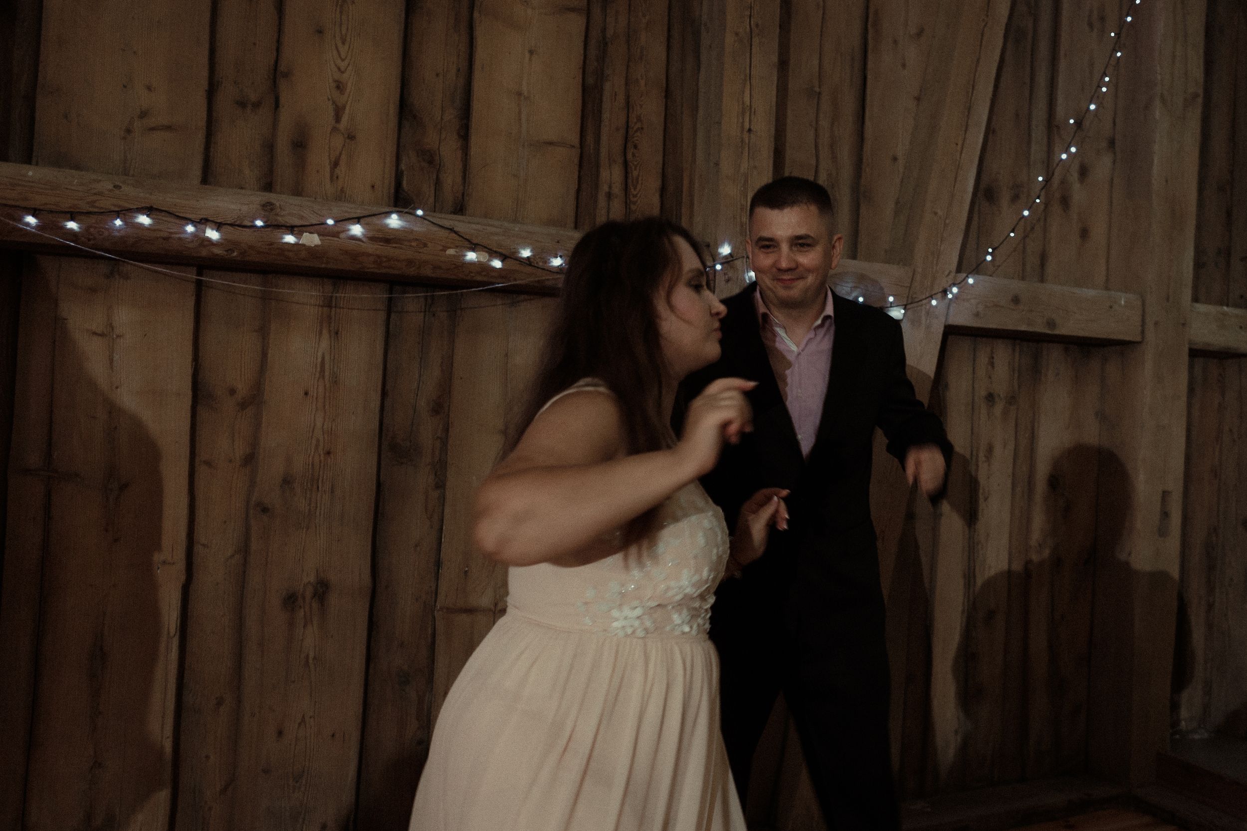 A group dances at a rustic barn reception with wooden paneled walls and festive string lights in the background.