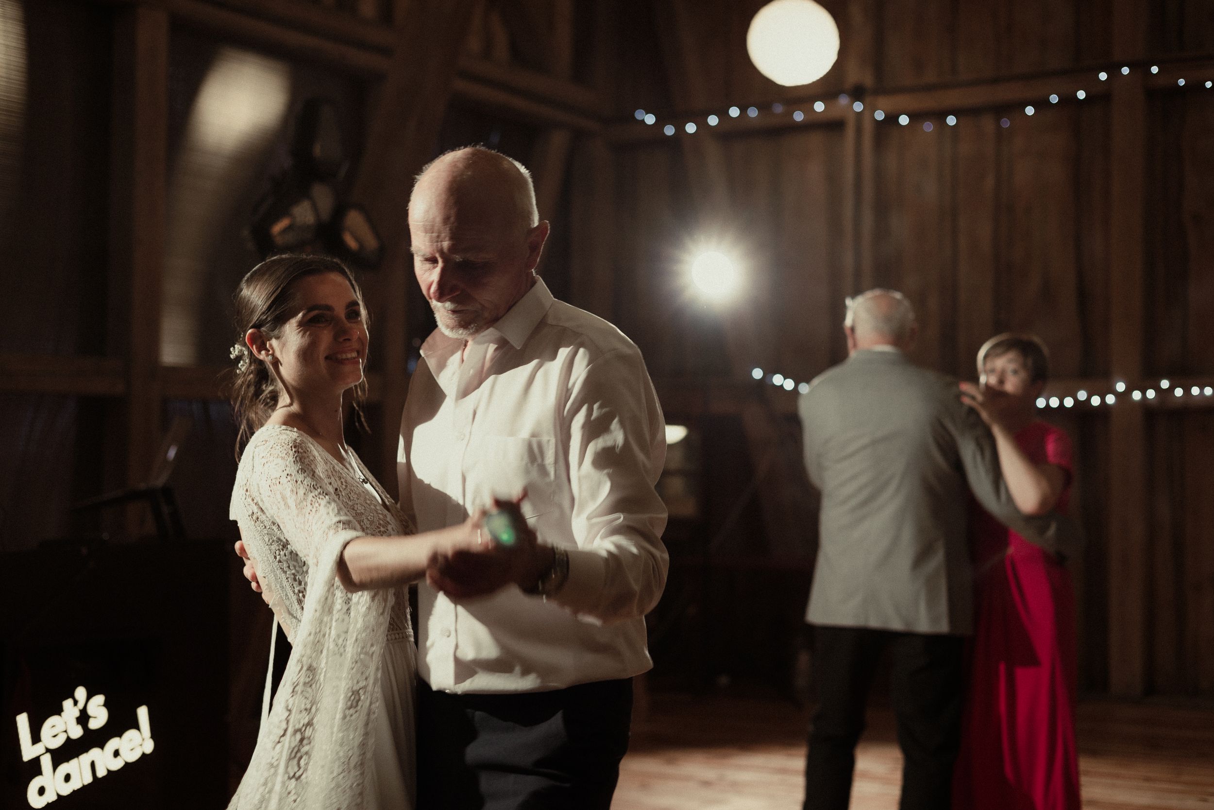 Couples slow dancing at an elegant indoor reception with string lights illuminating the rustic wooden beams above.