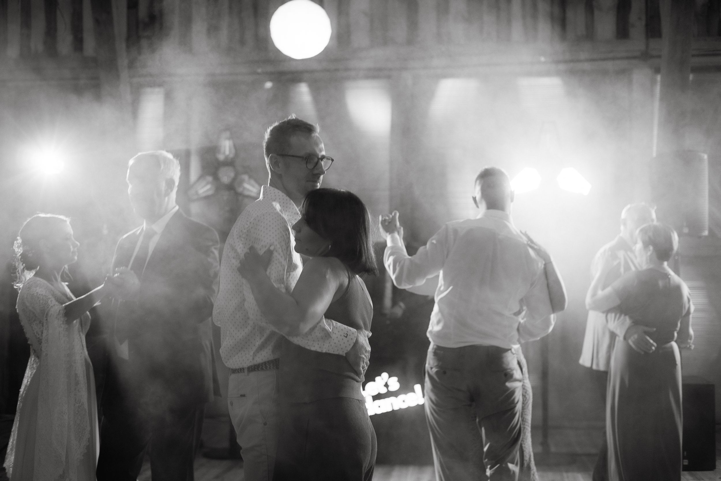 Black and white photo of silhouetted figures dancing at a wedding reception with bright lights in the background.