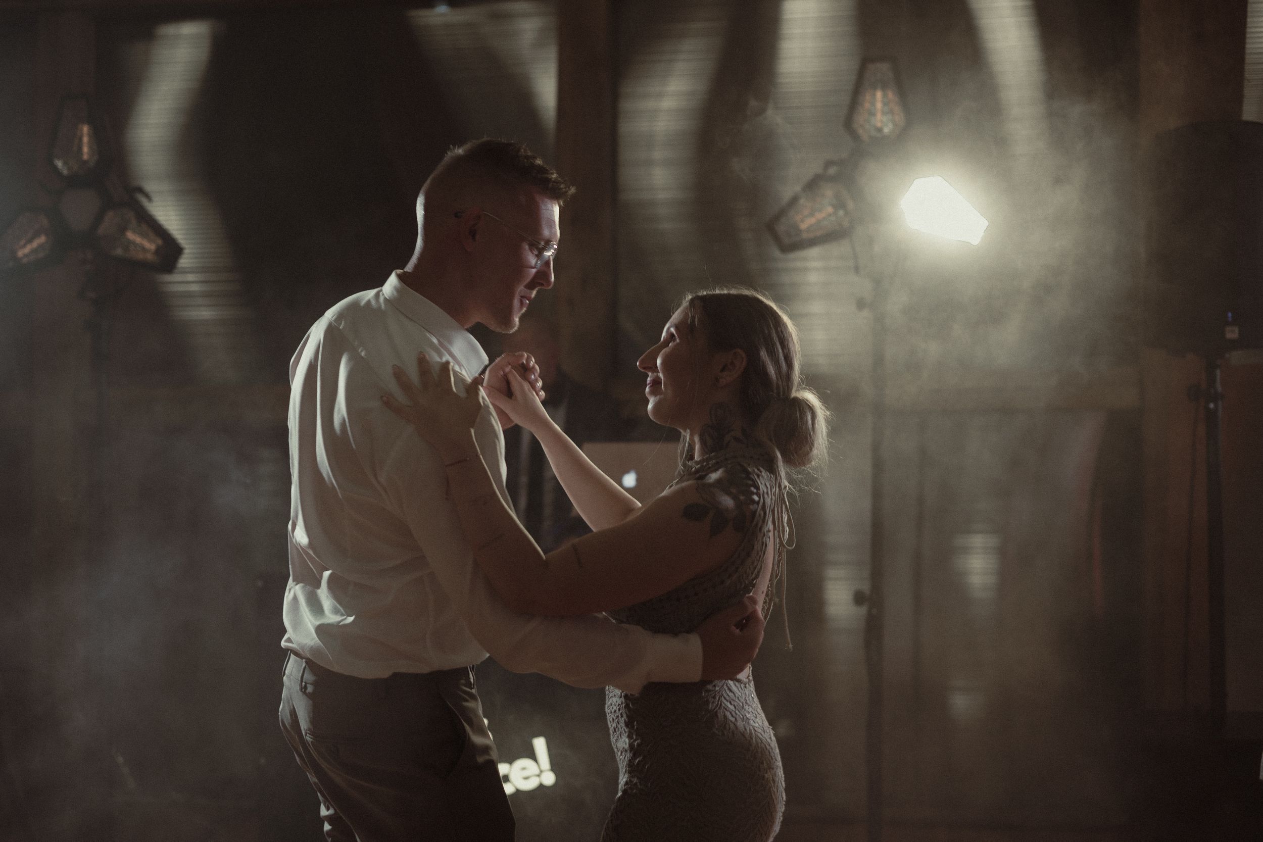 A romantic dancing couple share an intimate moment in a dimly lit industrial space with atmospheric fog and light beams.