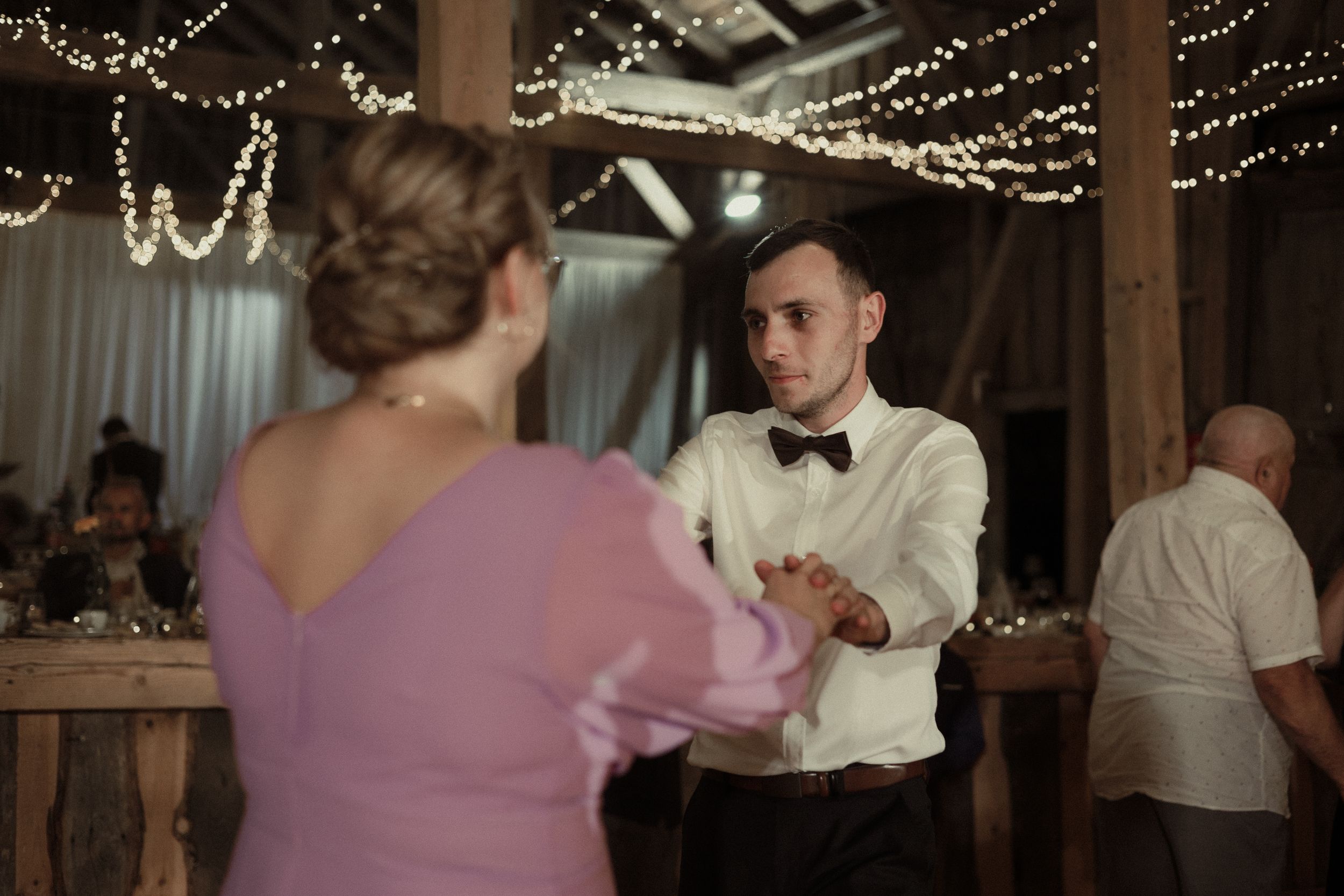A wedding reception dance party in a rustic barn venue decorated with string lights and featuring couples dancing.