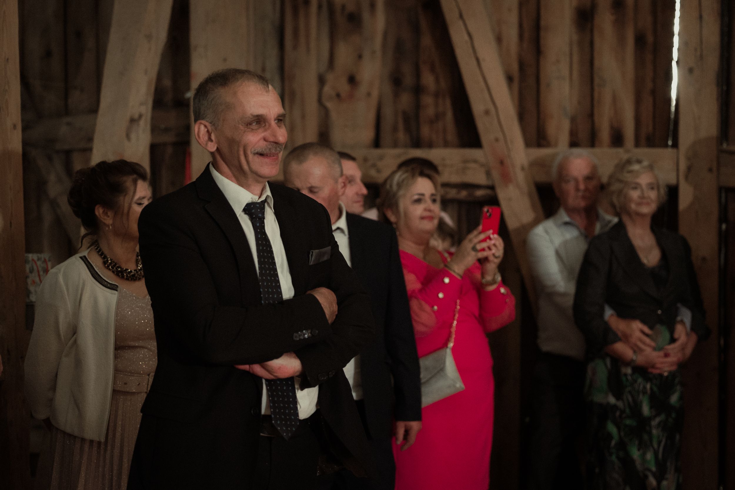 A group of formal event attendees laugh together inside a rustic wooden barn during a celebration.