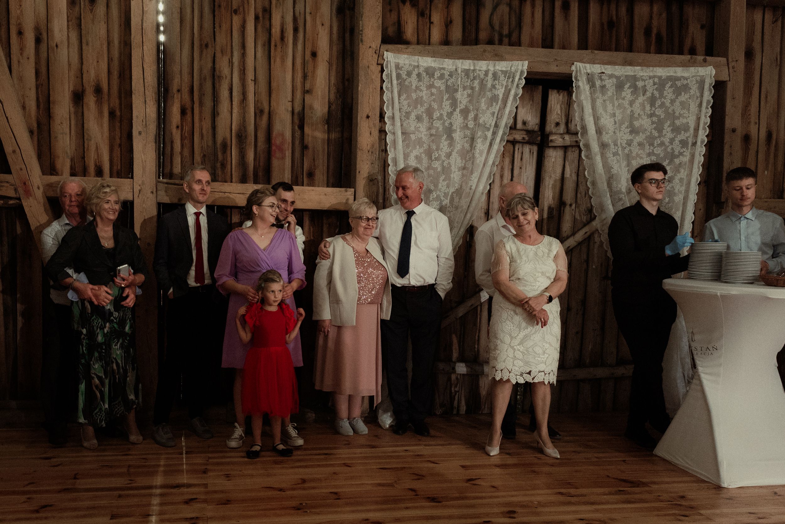 A group of wedding guests stands in line against a rustic barn wall with lace curtains and wooden details.