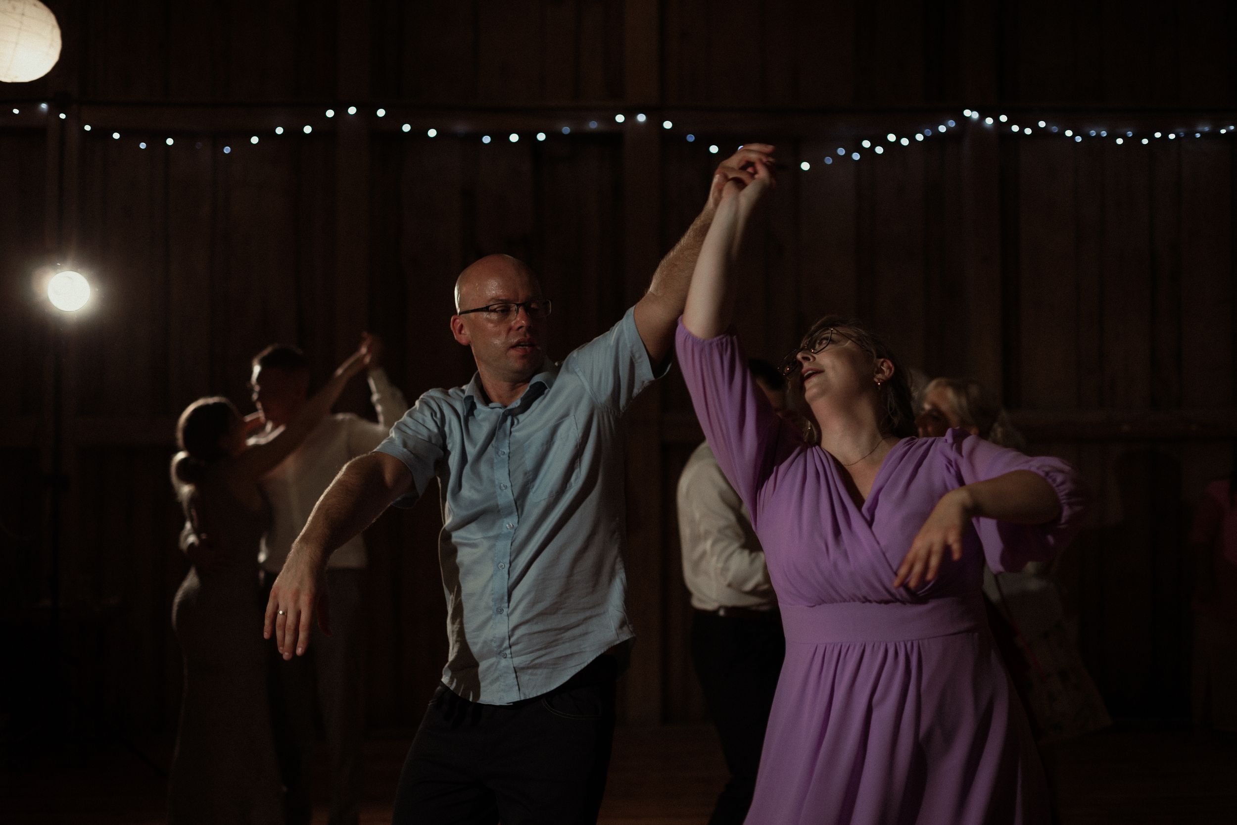 Couples dance together on a rustic wooden dance floor under dim lighting and string lights at an evening celebration.