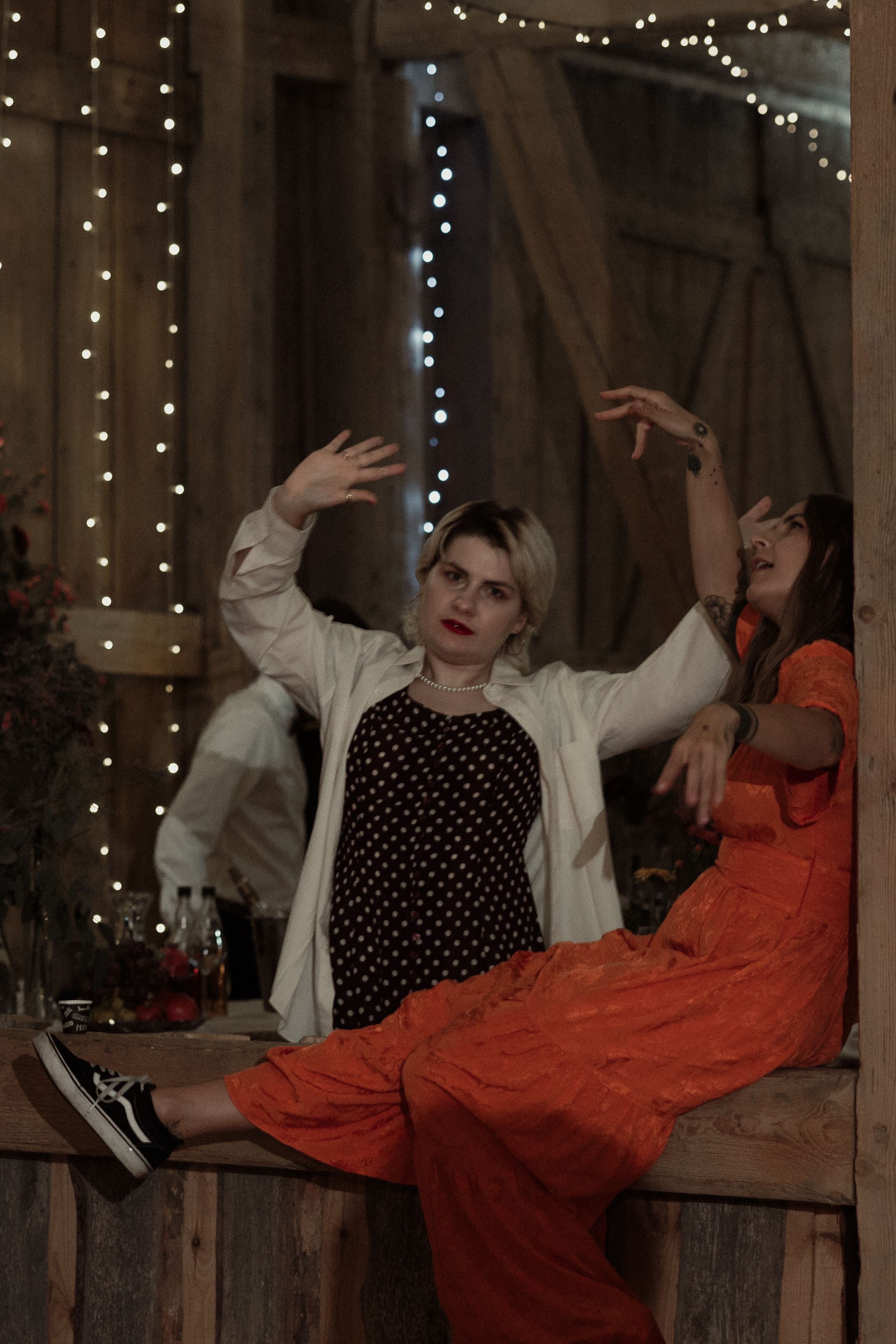 Two people in polka dot top and red dress pose casually on wooden porch with string lights in evening atmosphere.