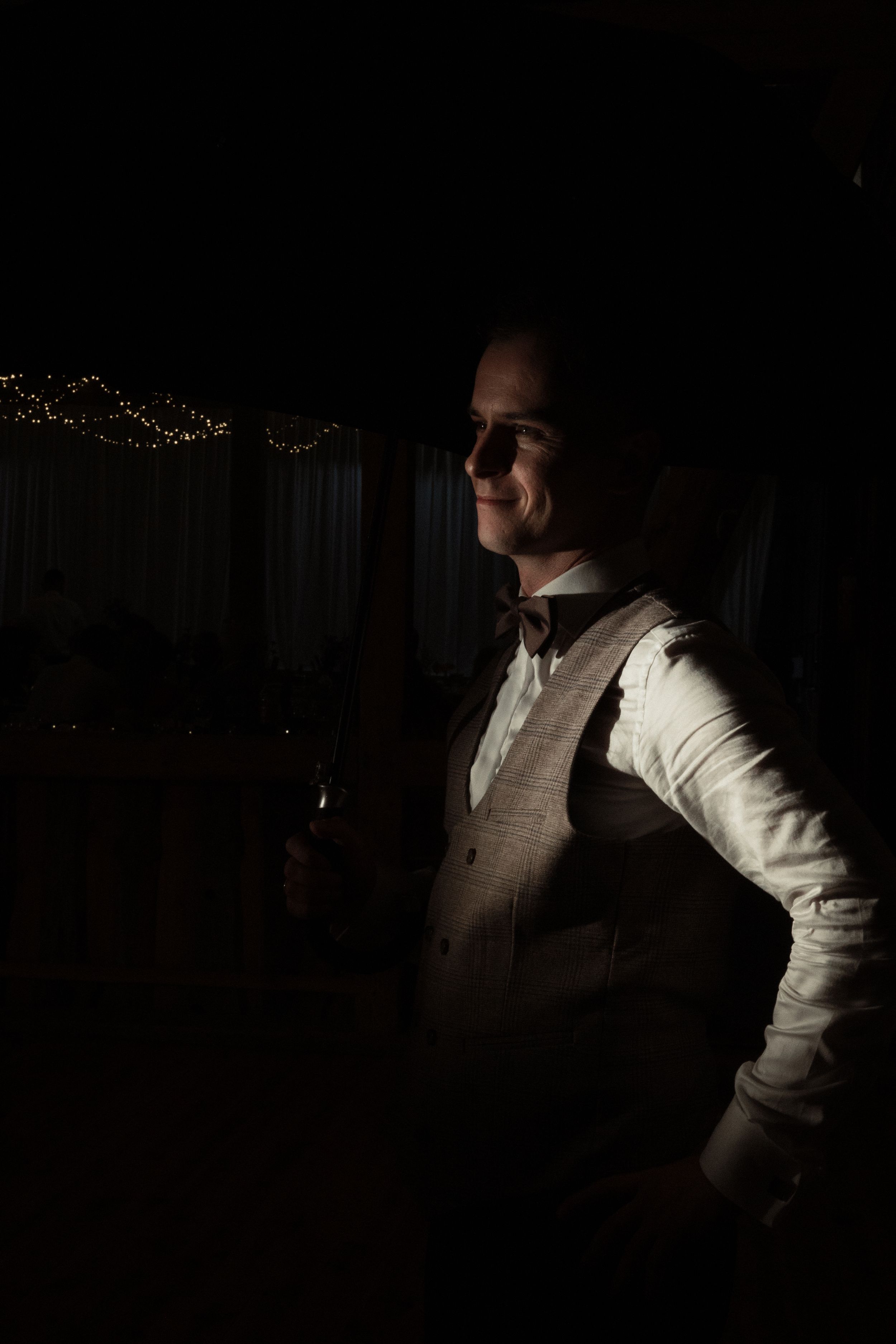 Someone in a white formal vest and dress shirt stands in a dark room with dramatic lighting casting shadows.