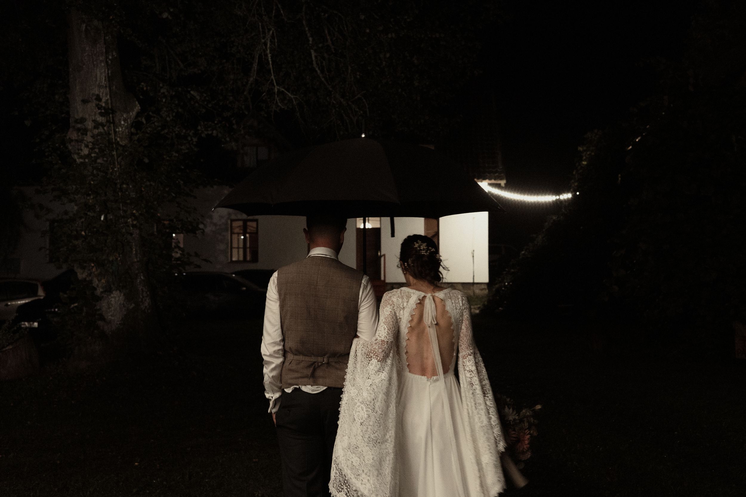 A bride in a white flowing dress holds a bouquet while standing in a dark nighttime outdoor venue.