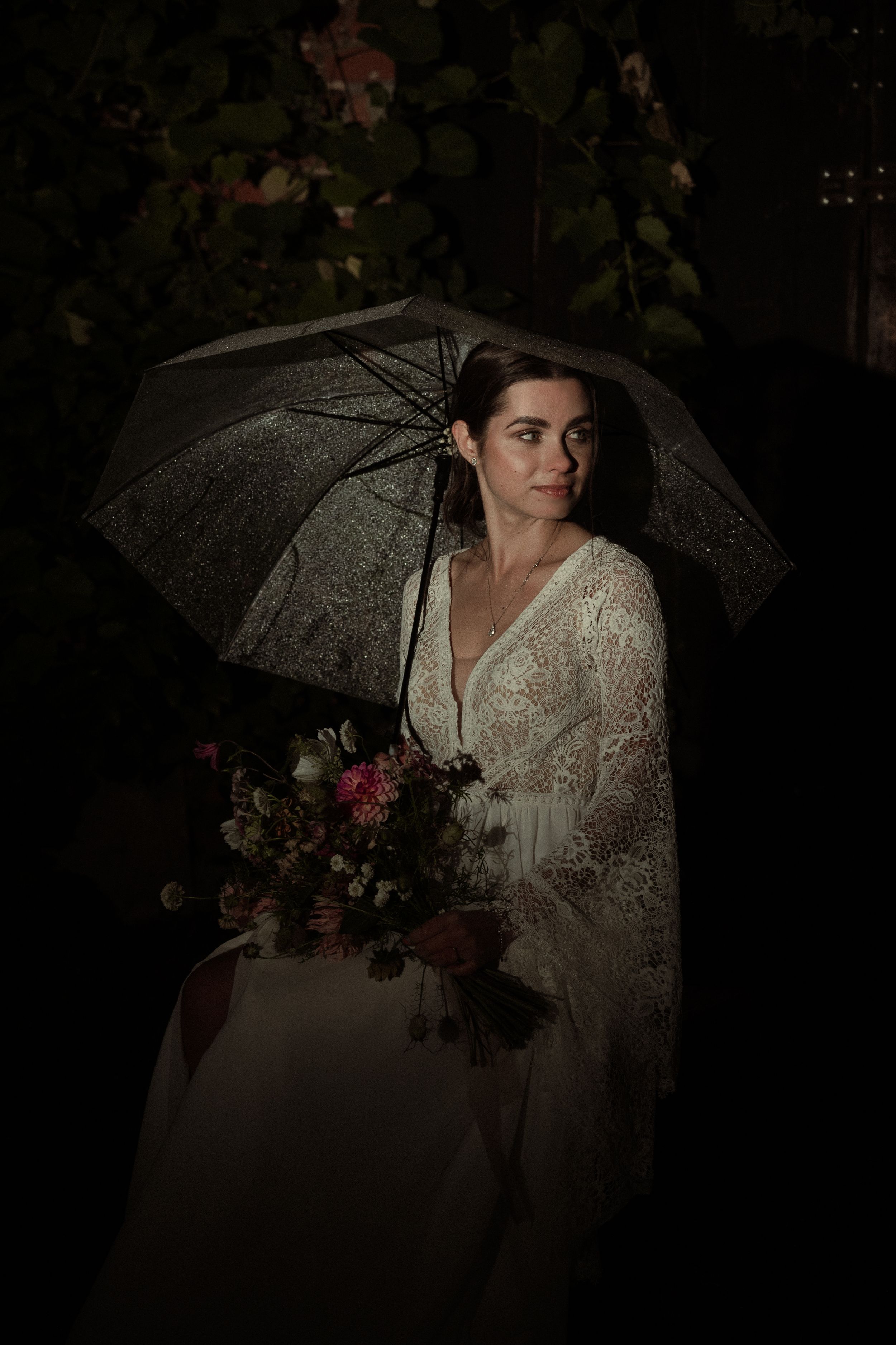 Moody wedding portrait with a white lace umbrella and bouquet against a dark background.