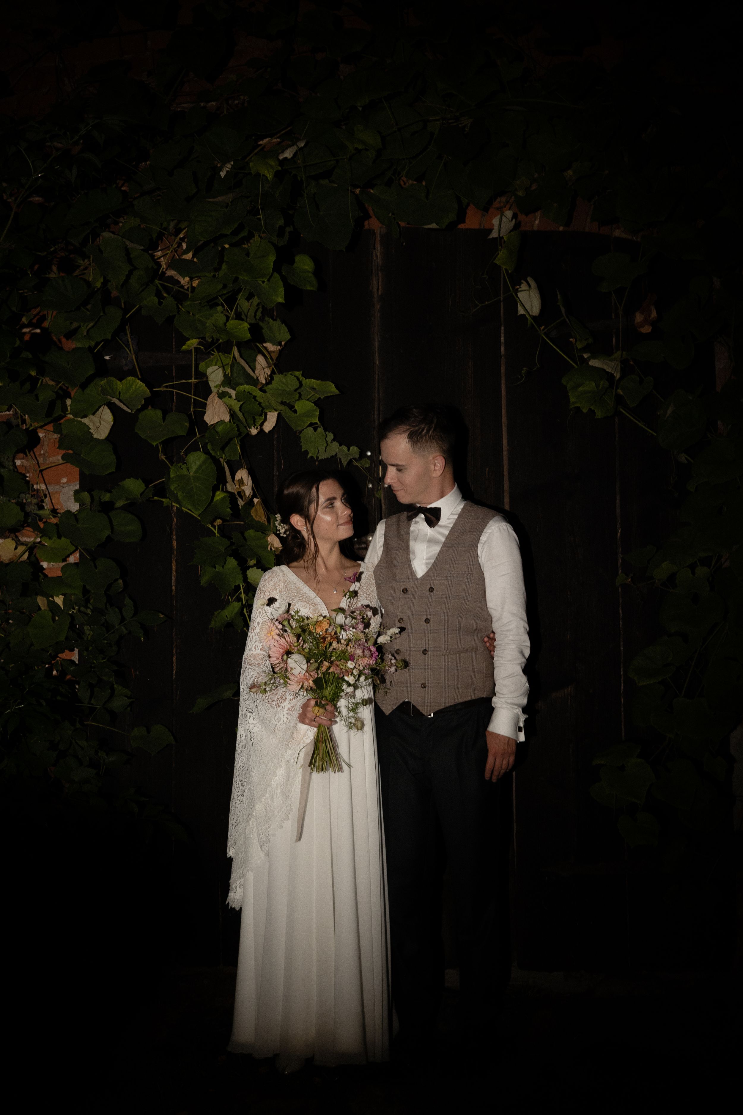 A romantic wedding couple poses outdoors at night against a dark background with subtle floral accents.