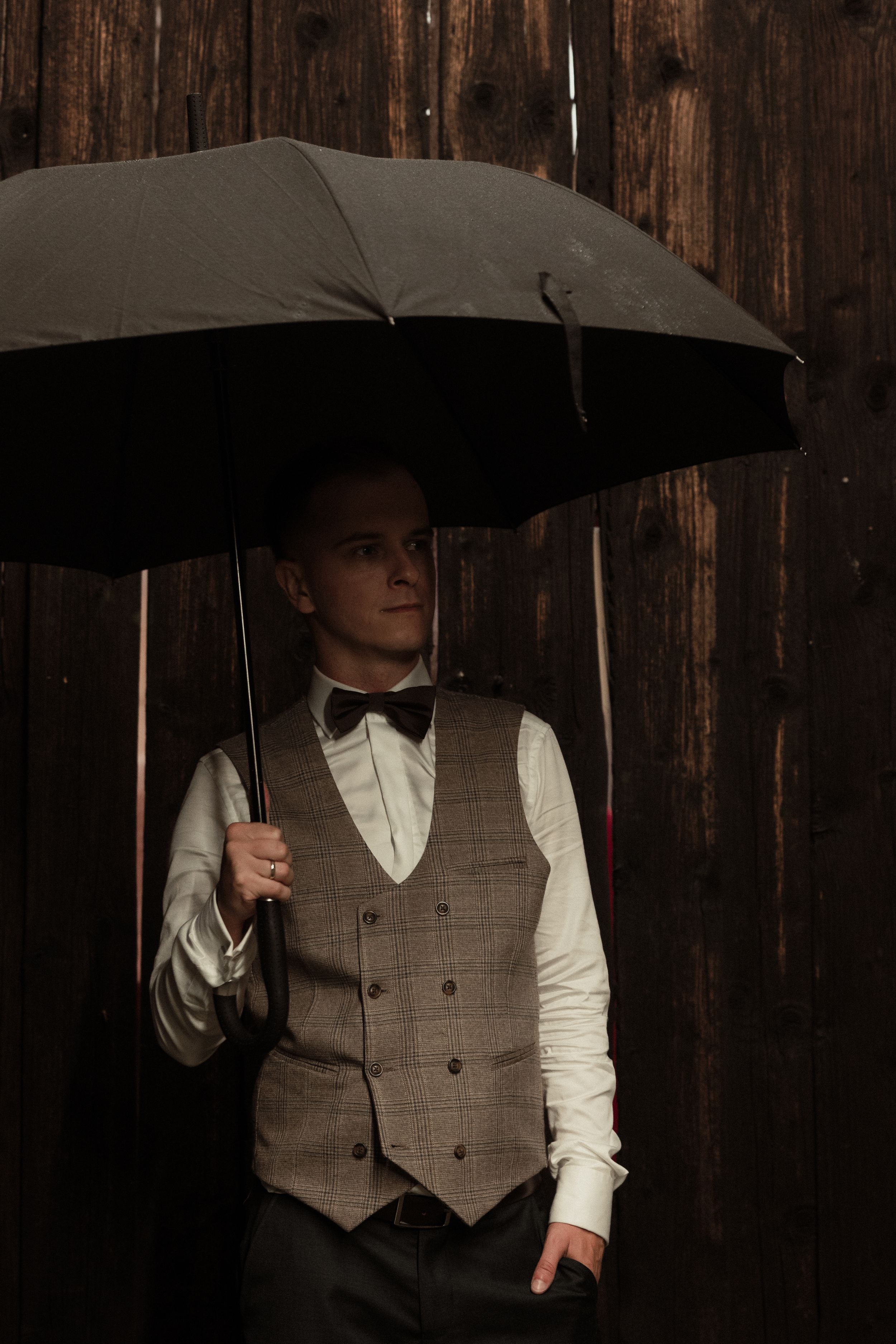 A figure in a beige vest and white shirt poses dramatically with a black umbrella against a dark wooden wall.