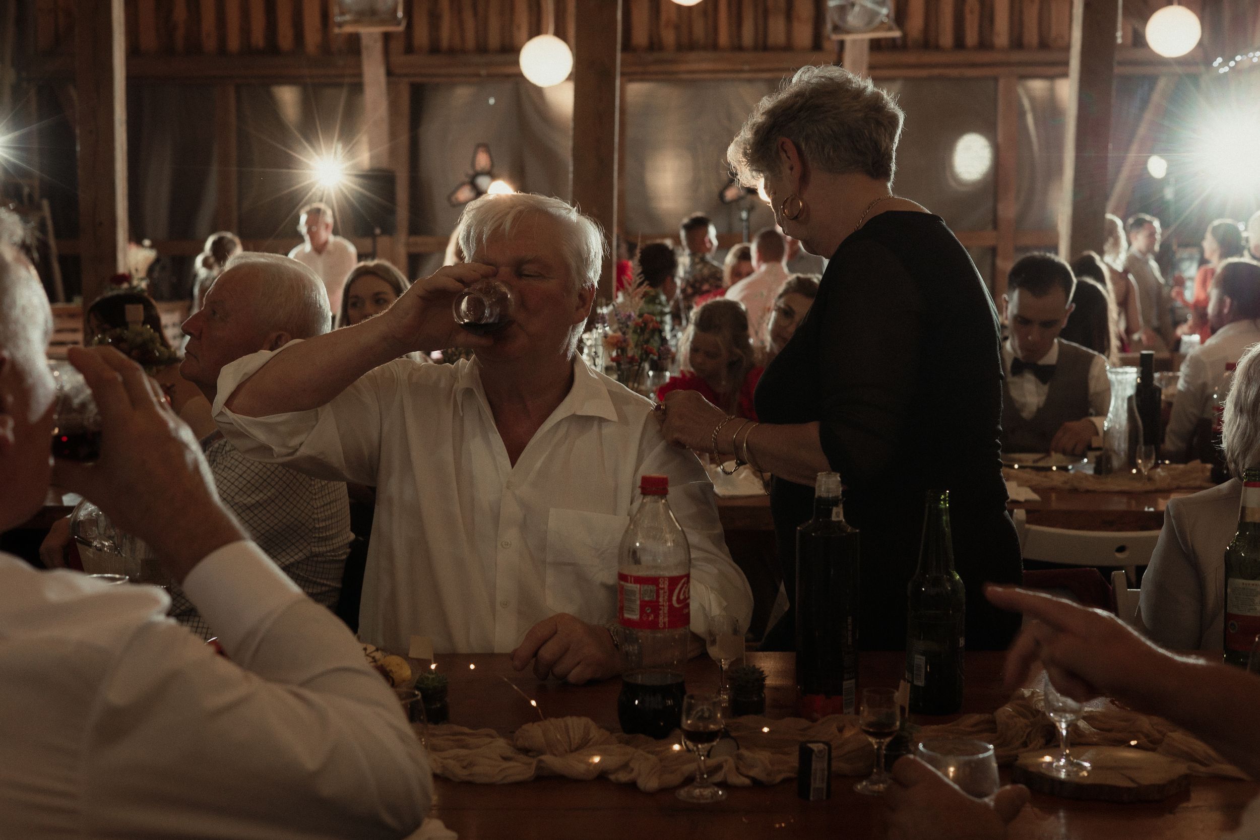 Wedding guests dancing at a dimly lit rustic barn reception with glowing string lights overhead.
