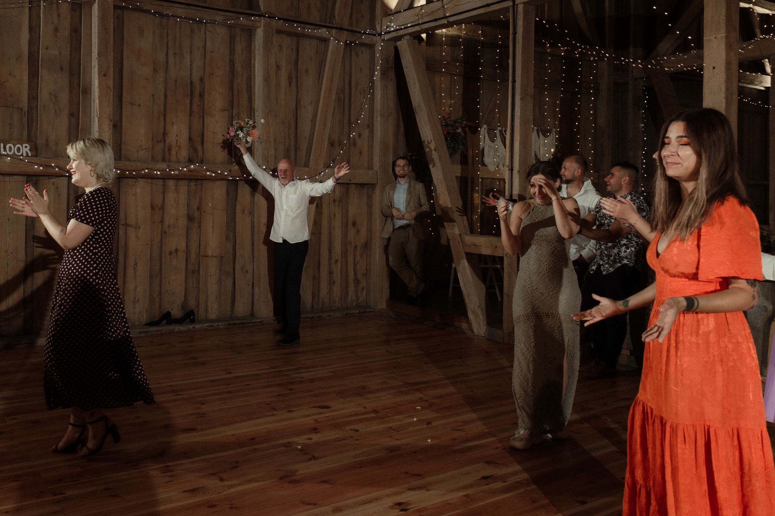 People dance and celebrate together at a rustic barn reception with string lights overhead.