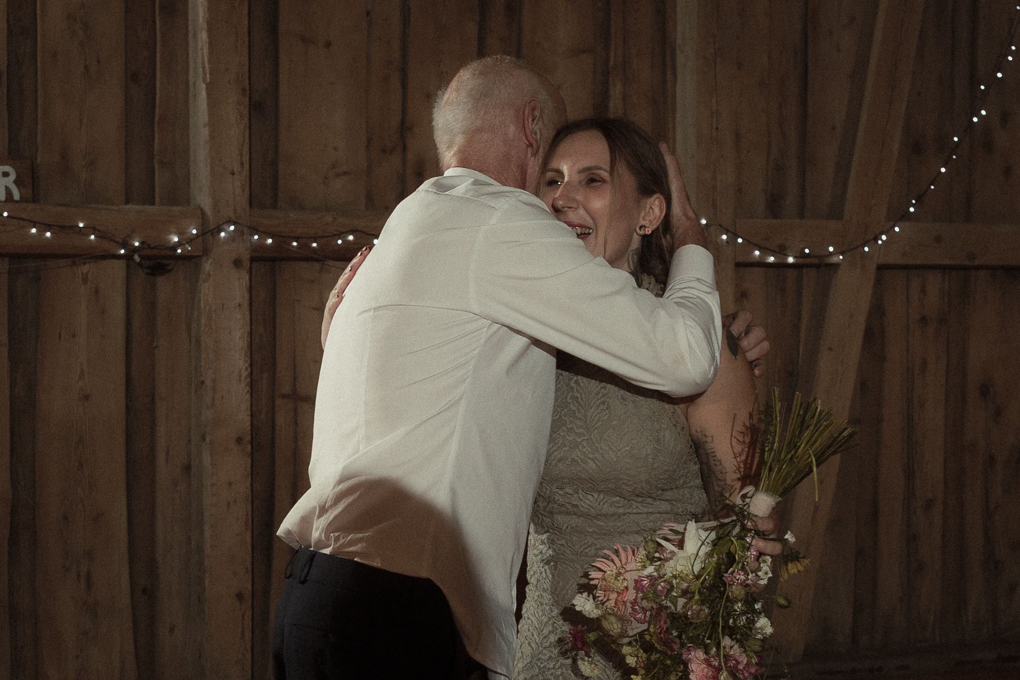 A romantic moment in a rustic barn venue with string lights and floral decorations during a wedding celebration.