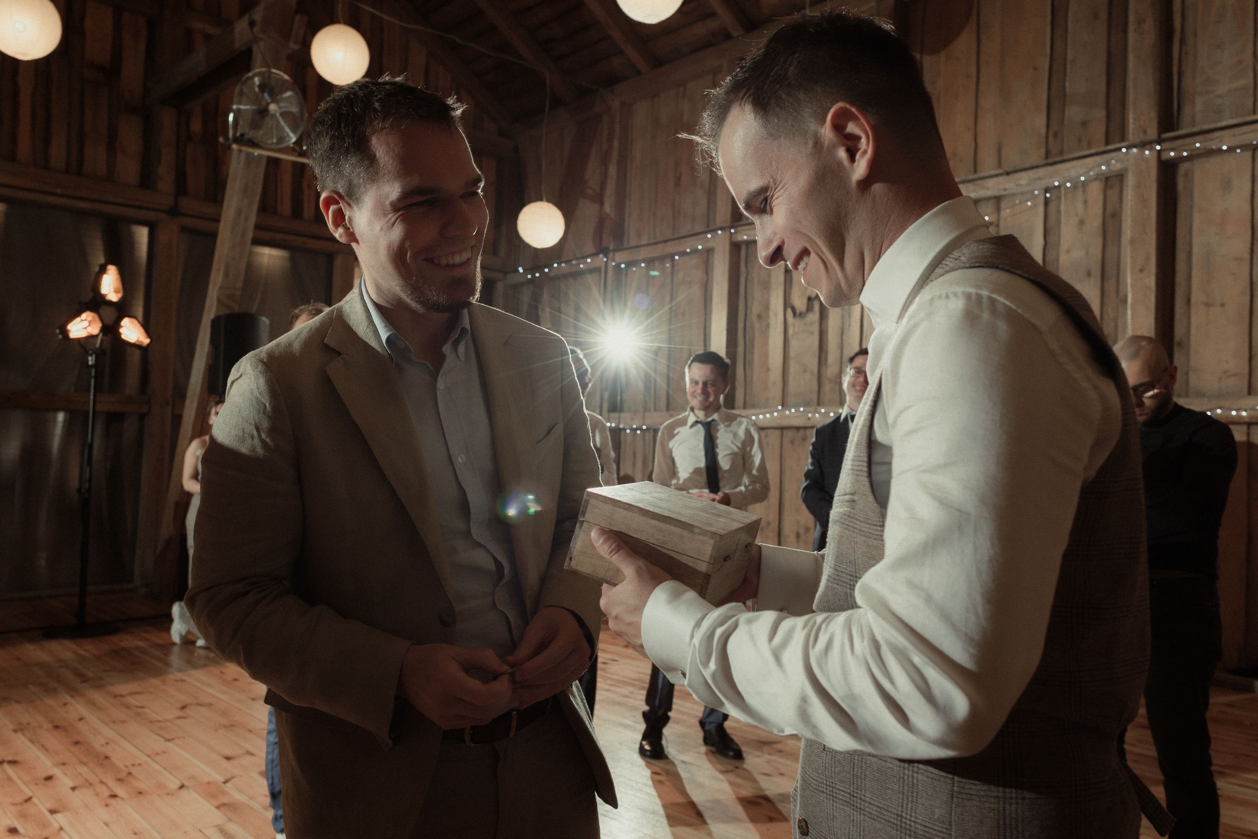 Two people share a joyful moment while reading from papers in a rustic barn venue with glowing string lights overhead.
