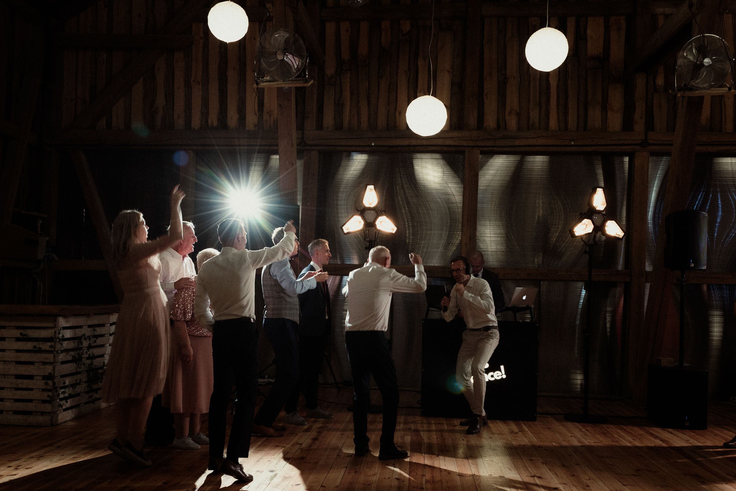 Wedding guests dance together under glowing spherical lights in a rustic wooden barn venue during an evening reception.