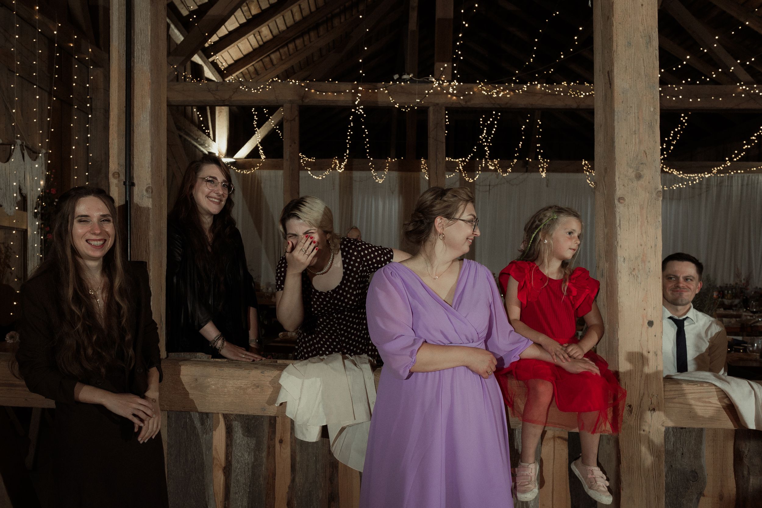 Guests dancing together joyfully at an indoor wedding reception with string lights and rustic wooden beams overhead.