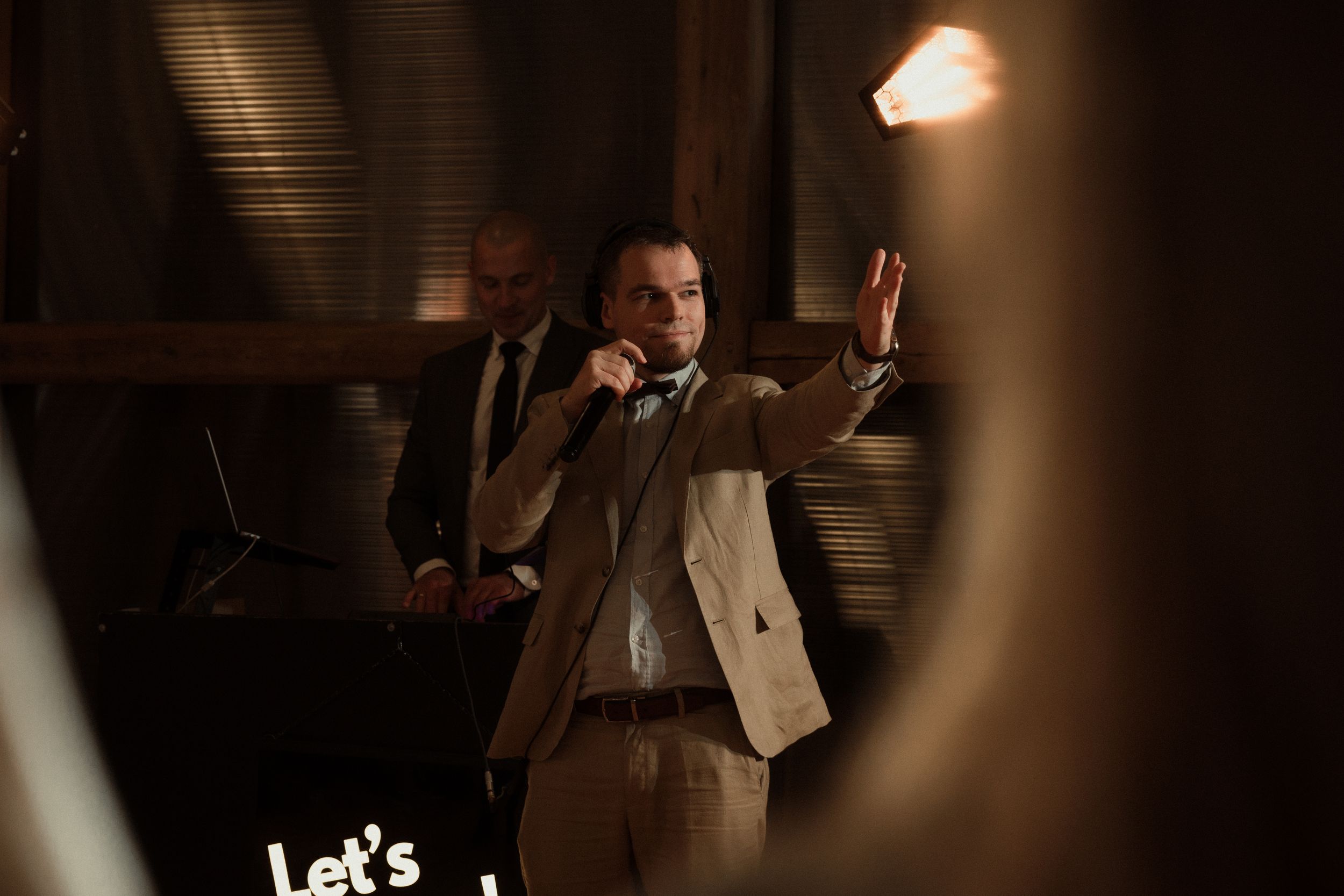 Person in brown suit and white jacket gestures while speaking in a dimly lit room with dramatic lighting.