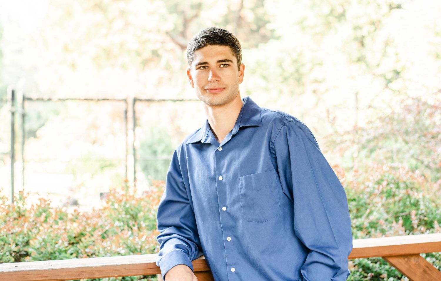 Person in blue button-down shirt leans against wooden railing during outdoor photo session.