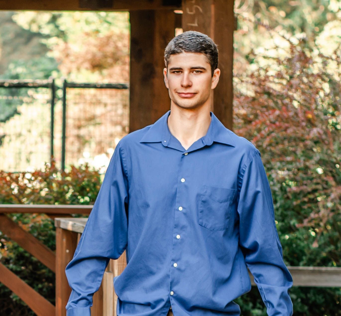 Person in blue button-down shirt poses against wooden railings in outdoor setting.