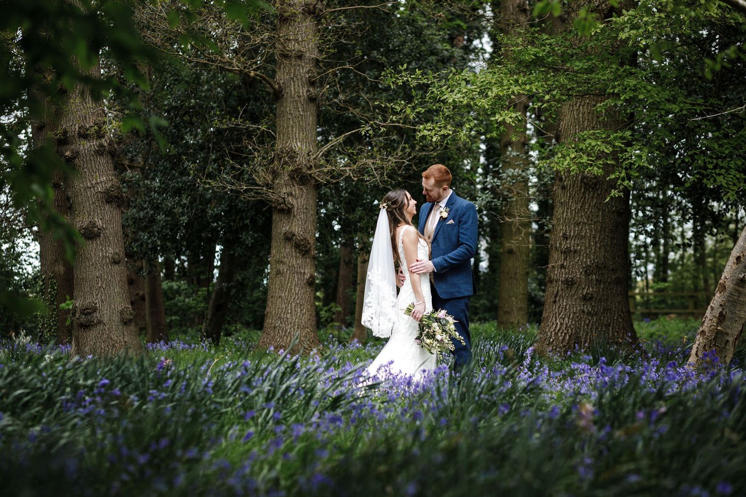 Newlywed couple sharing a tender moment in the Kelham House woodland surrounded by purple bluebells beneath tall trees