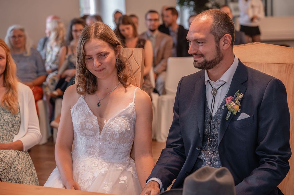 A wedding couple sits together during their ceremony while guests look on in a bright indoor venue.