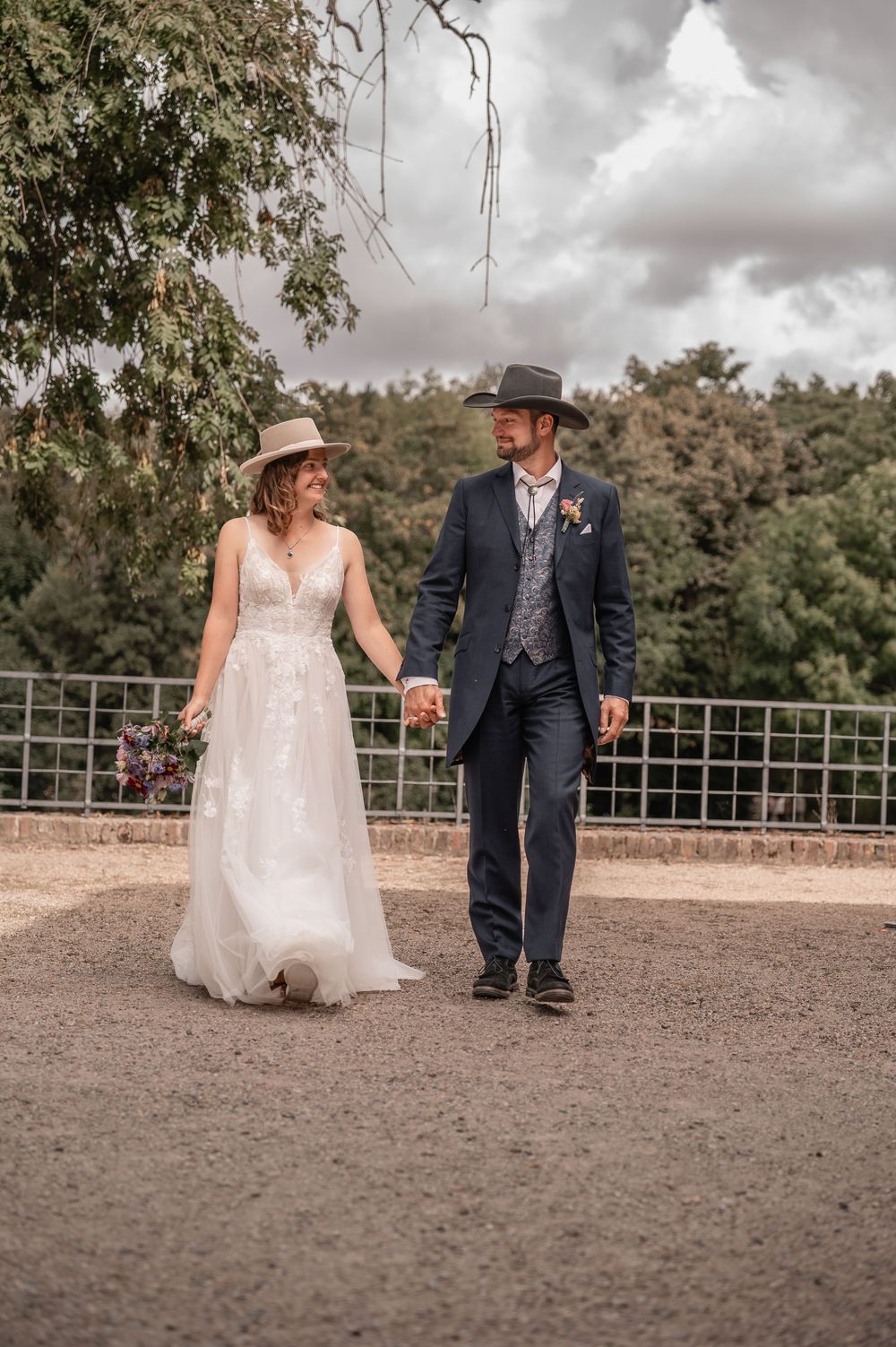 A couple walks hand in hand along a scenic path, the bride in white and groom in navy with western-style hats.