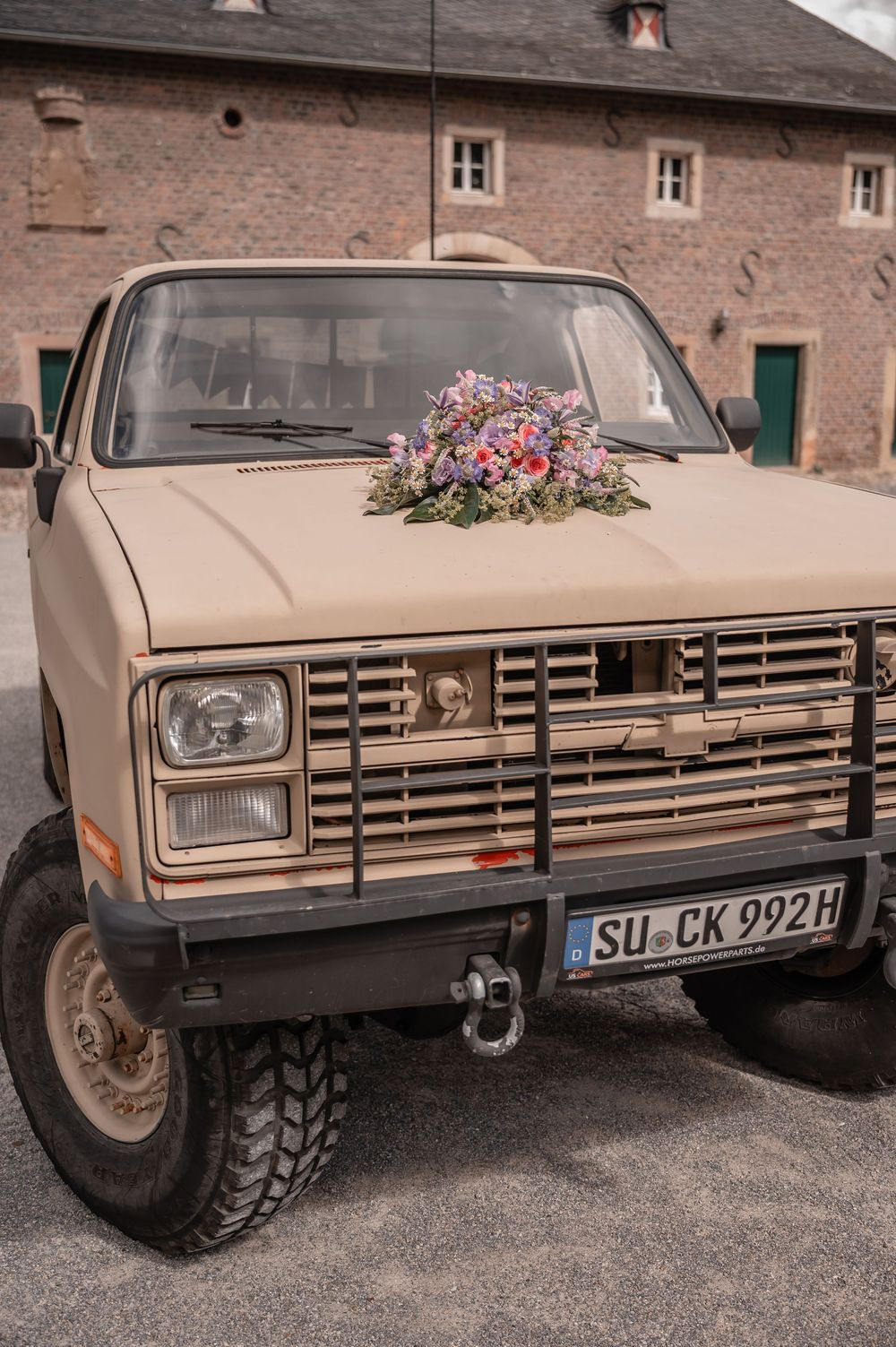 Vintage pickup truck with floral decorations on its front grille parked against a rustic brick building backdrop.