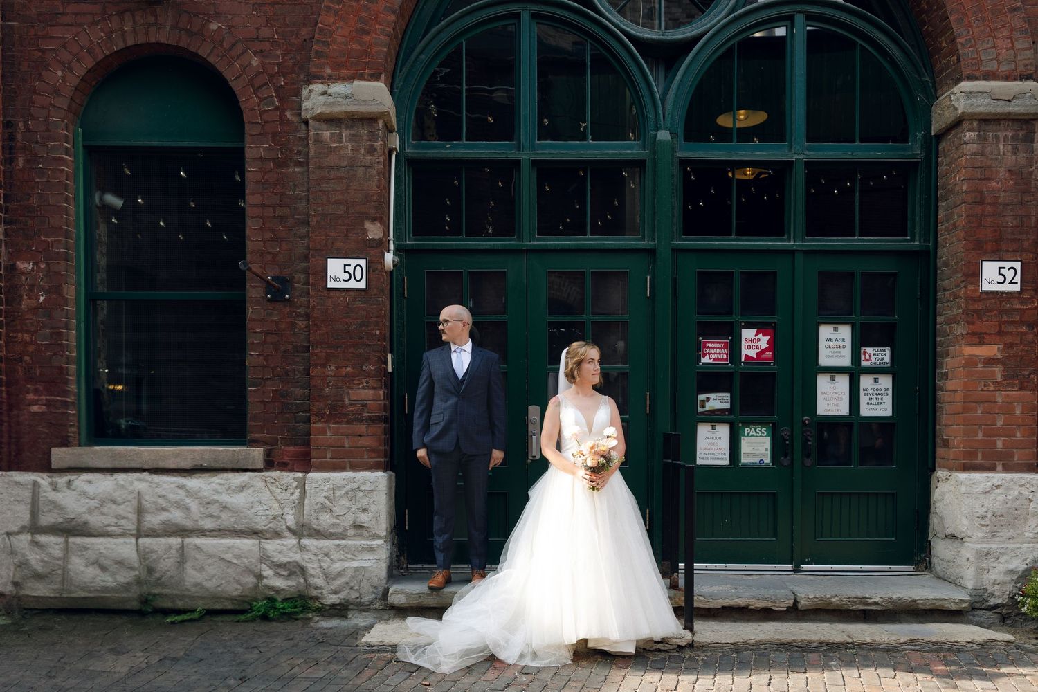 A couple in wedding attire walk hand in hand along a rustic brick-paved path beside historic industrial buildings.