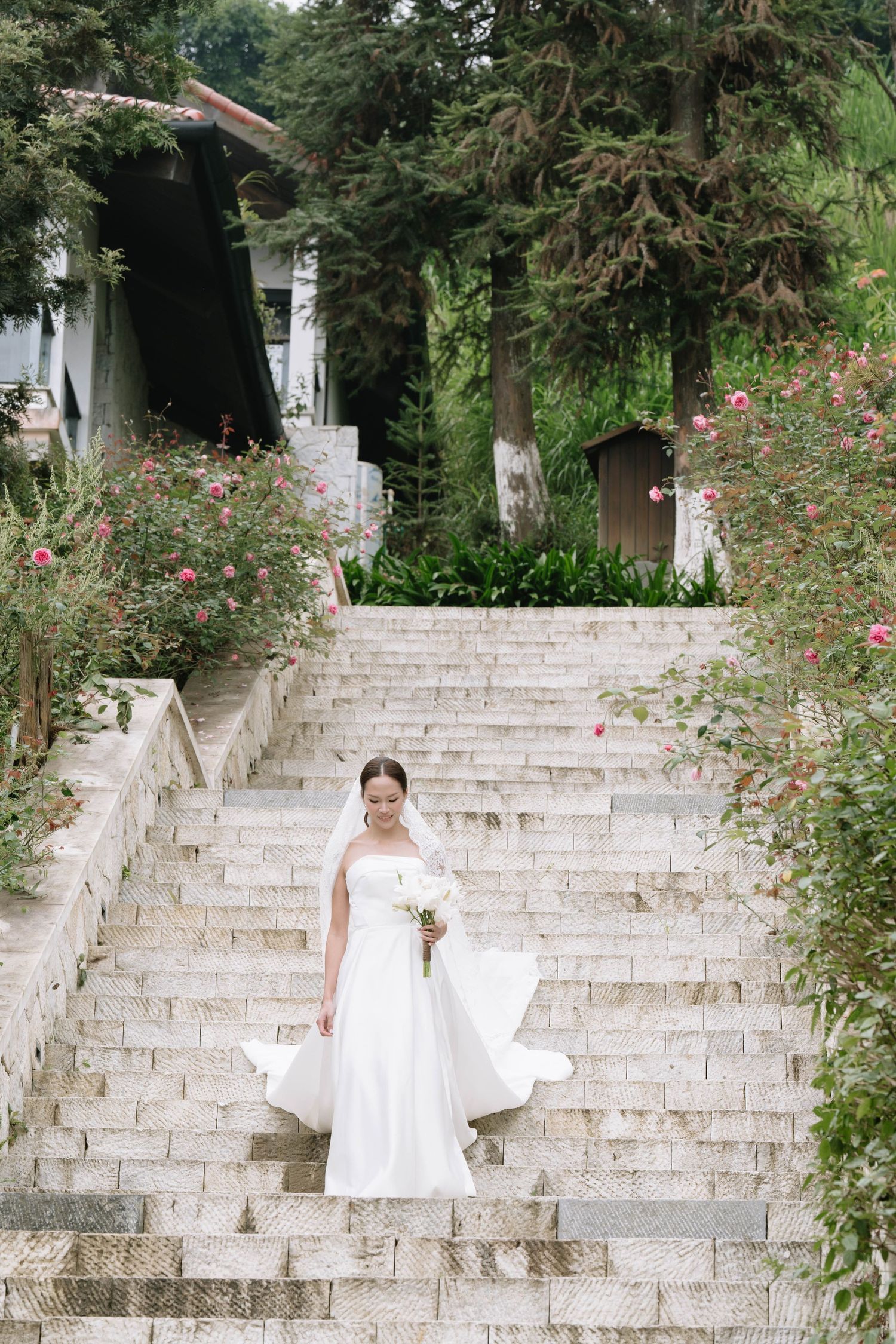 Bride in flowing white strapless gown descends grand stone staircase surrounded by lush greenery.