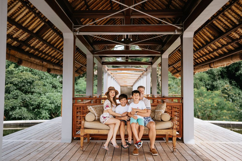 Family relaxing together on a wooden bench in a covered tropical pavilion.