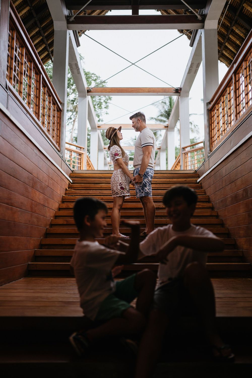 Family members share playful moments on wooden stairs in a tropical resort setting.