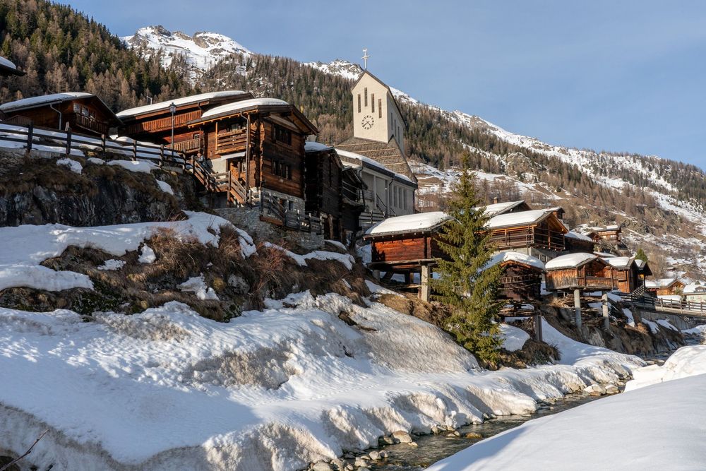 A panoramic winter view of a snow-covered alpine village nestled in the mountains with wooden chalets and ski slopes.