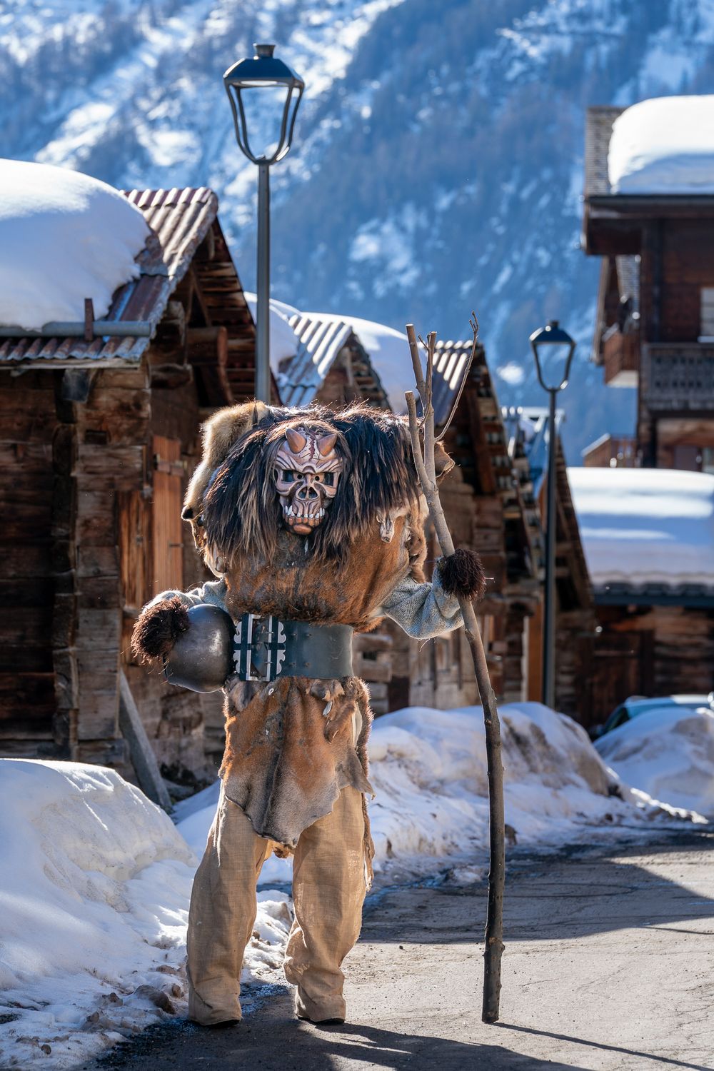 A creepy scarecrow-like figure wearing ragged clothes stands in a snowy mountain landscape near a street lamp.
