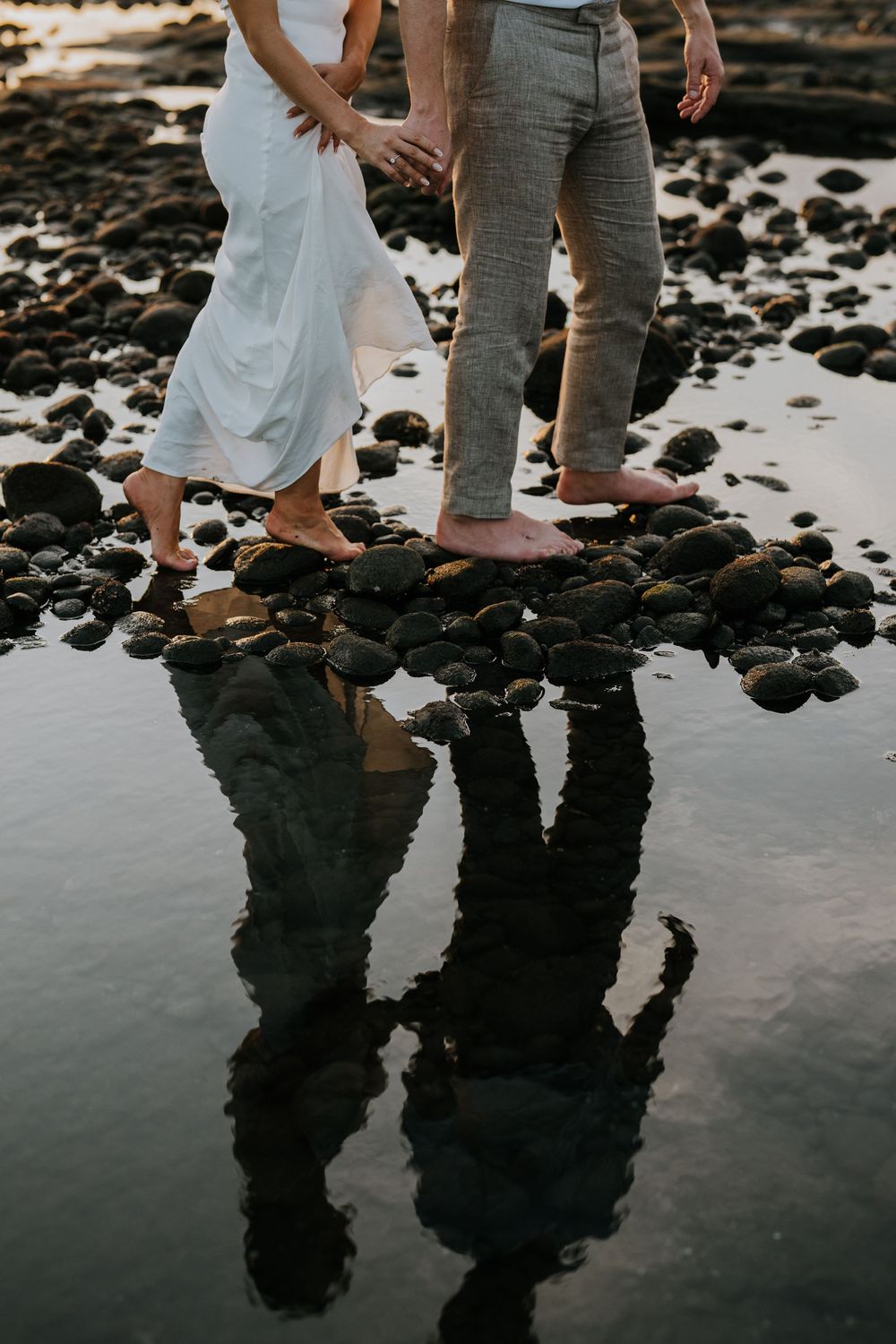 Couple's legs and reflections on wet rocky beach at sunset creating mirror effect in tide pools.
