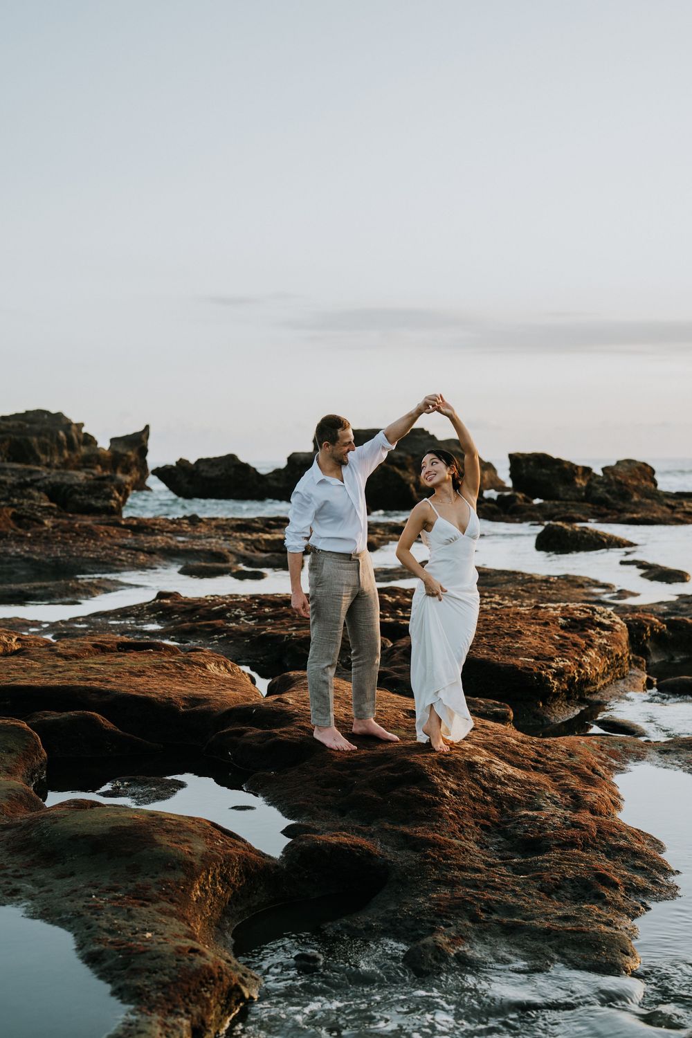 Couple dancing on rocky coastal outcrop during sunset with ocean waves below.