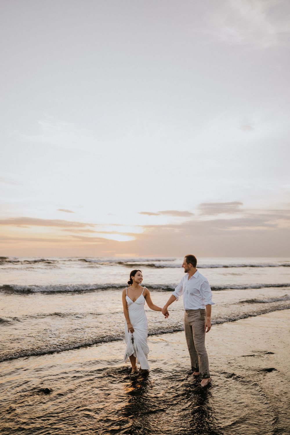 Couple walking hand in hand along shoreline during golden hour with waves rolling in.