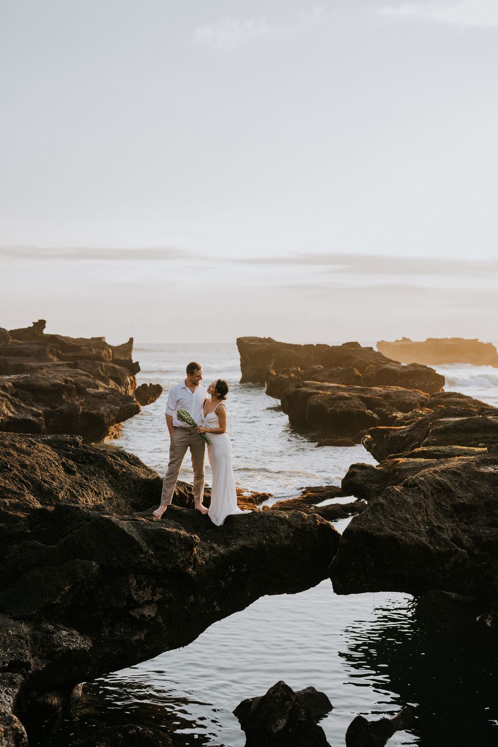 A couple standing on dramatic coastal rocks overlooking the ocean at sunset.