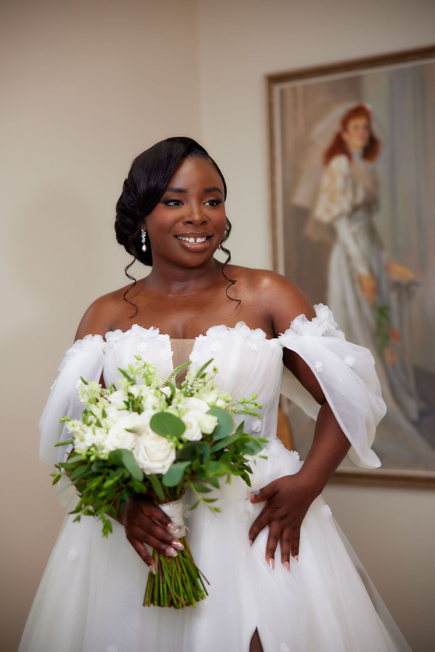 A bride in a white off-shoulder wedding gown holds a beautiful white and green bouquet against an indoor backdrop.