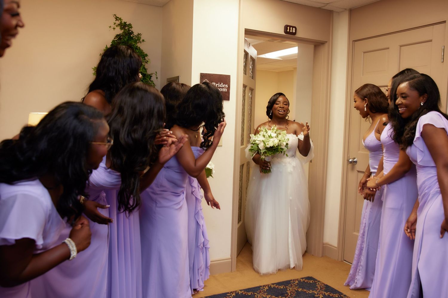 People in lavender bridesmaid dresses react with excitement as bride in white gown appears in doorway.
