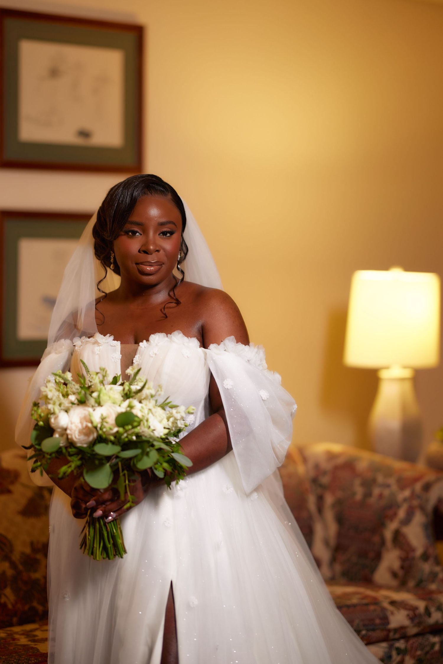A bride in a white wedding gown holds a lush bouquet of white flowers and greenery against a warm beige interior setting.