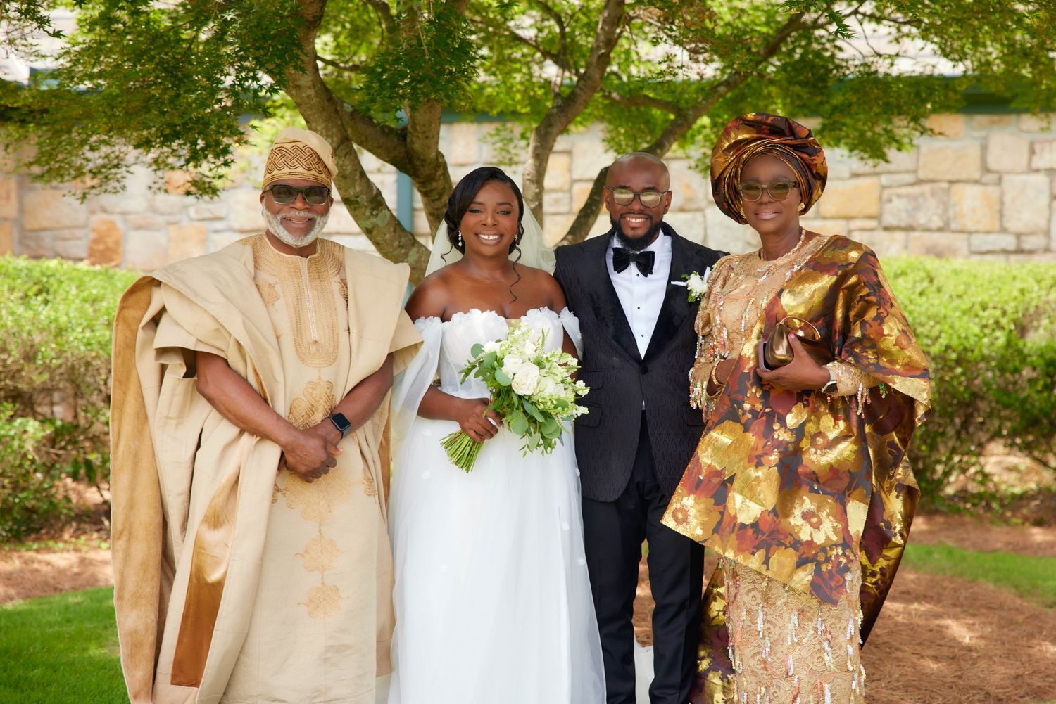 A wedding party in traditional African attire poses together outdoors with the bride wearing a white gown and groom in tuxedo.