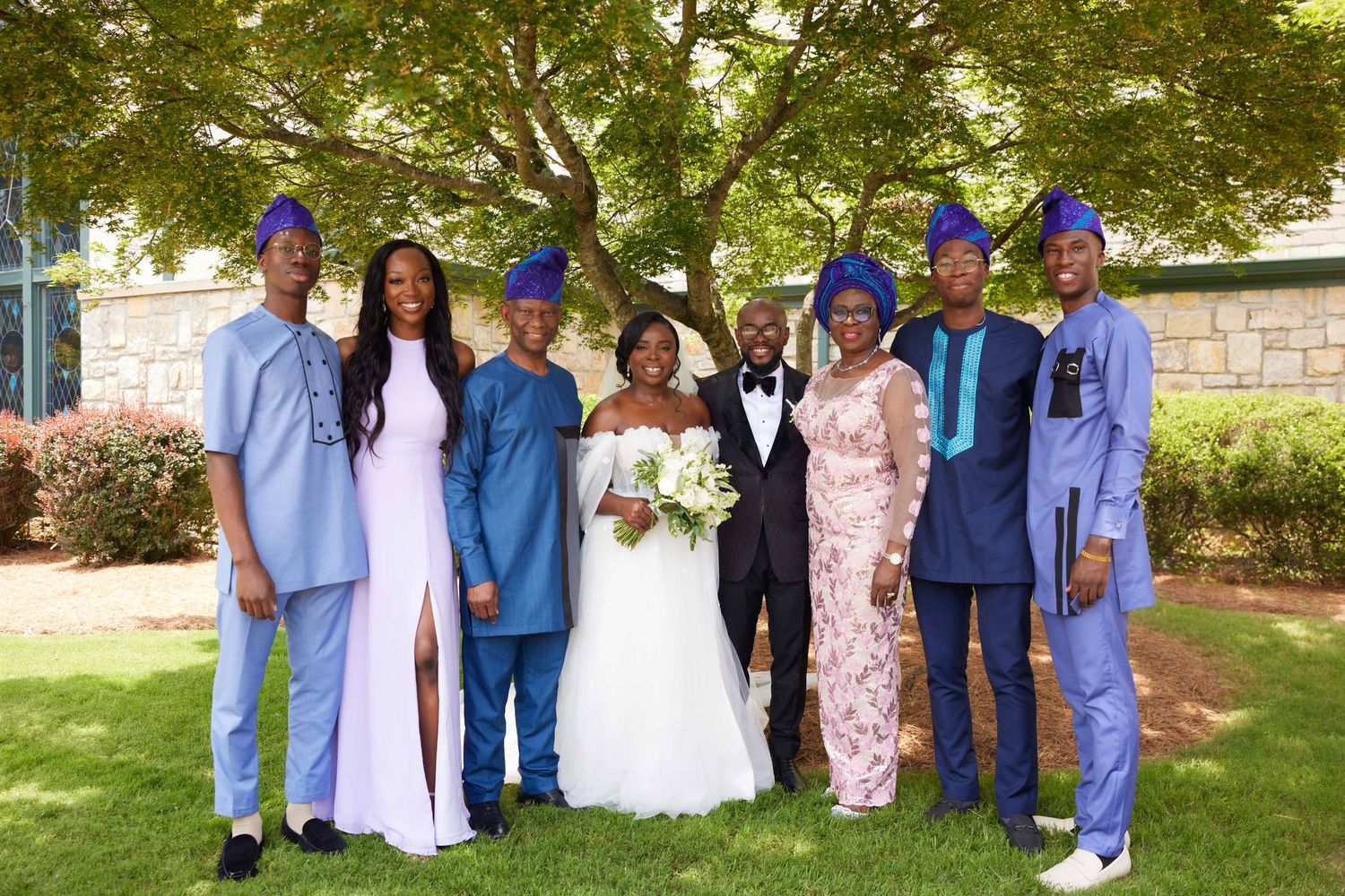 Wedding party in coordinating blue and pink attire poses together on a sunny lawn during outdoor celebration.