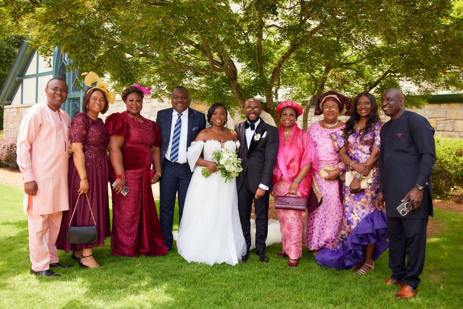 A wedding party in pink and burgundy attire poses together outdoors under green trees.