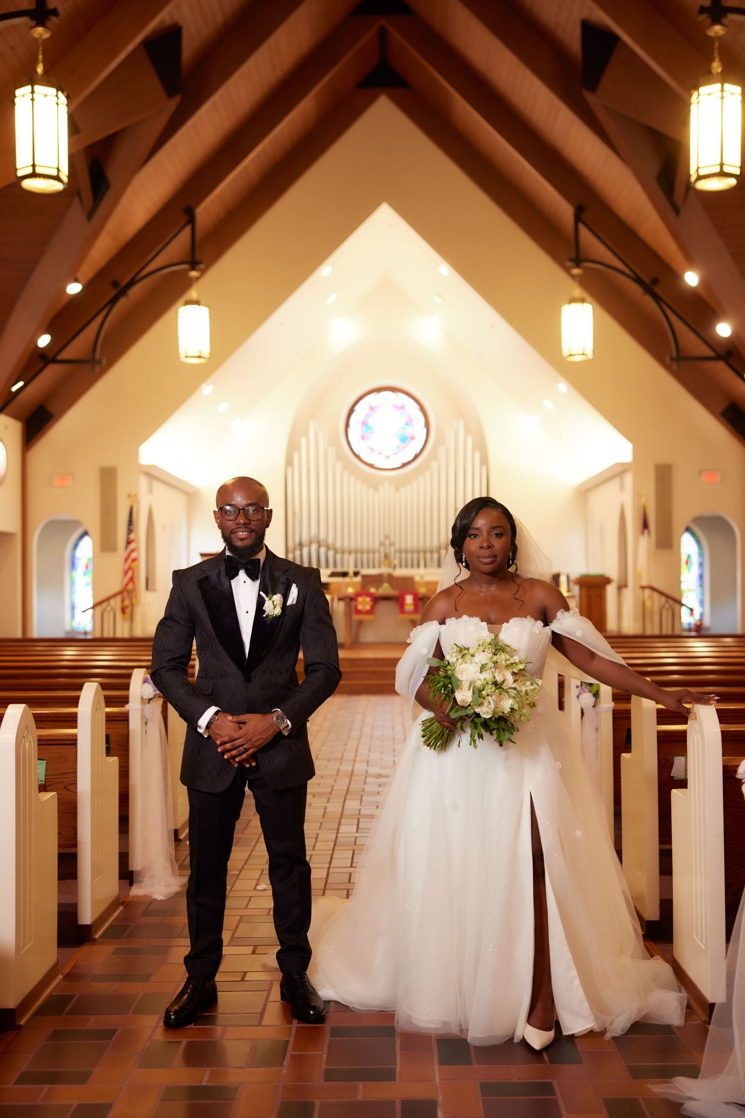A couple stands in a church aisle with vaulted ceilings, pendant lights and wooden pews as backdrop for their wedding ceremony.