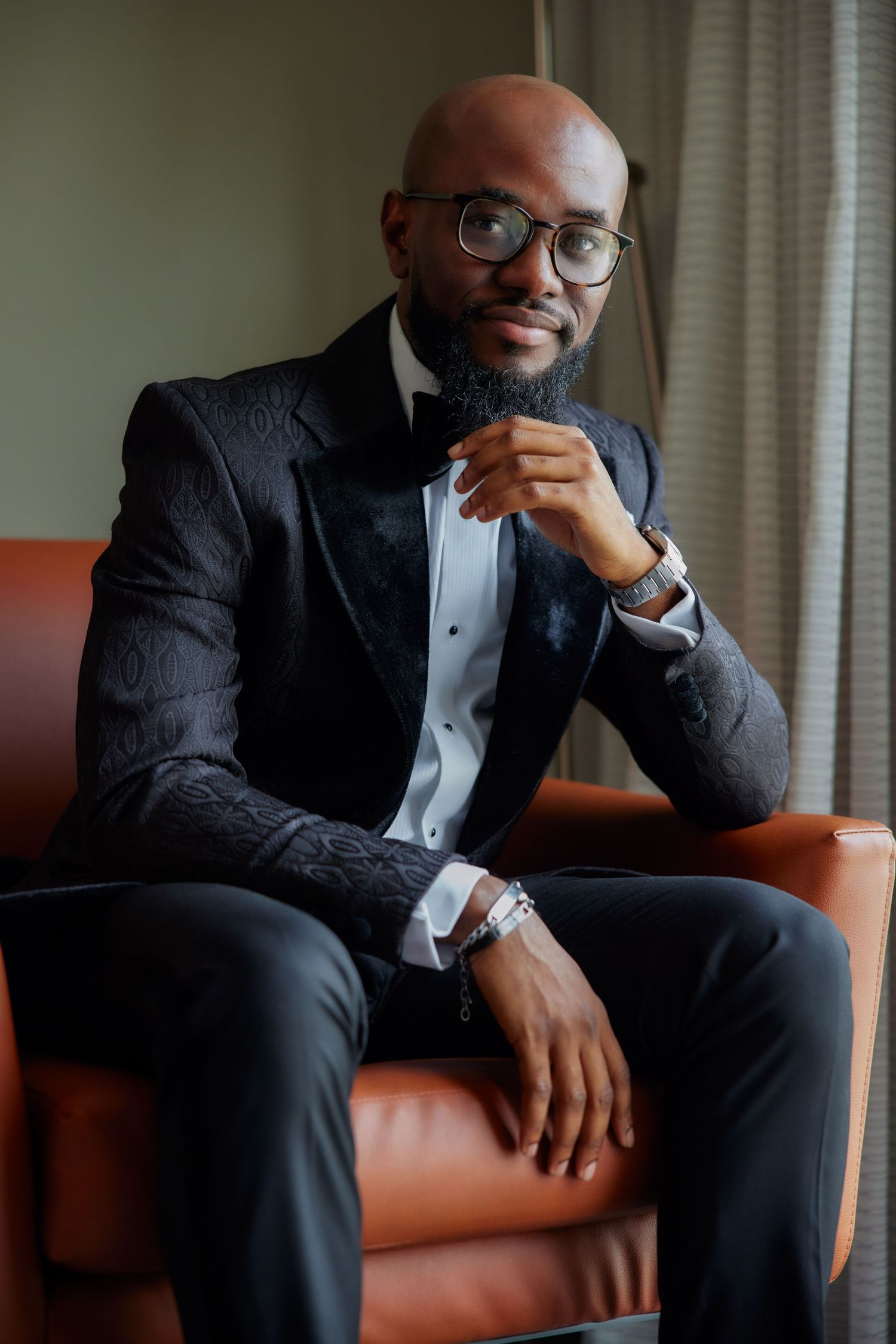 A person in a black tuxedo and glasses sits thoughtfully on a brown leather couch in multiple similar poses.