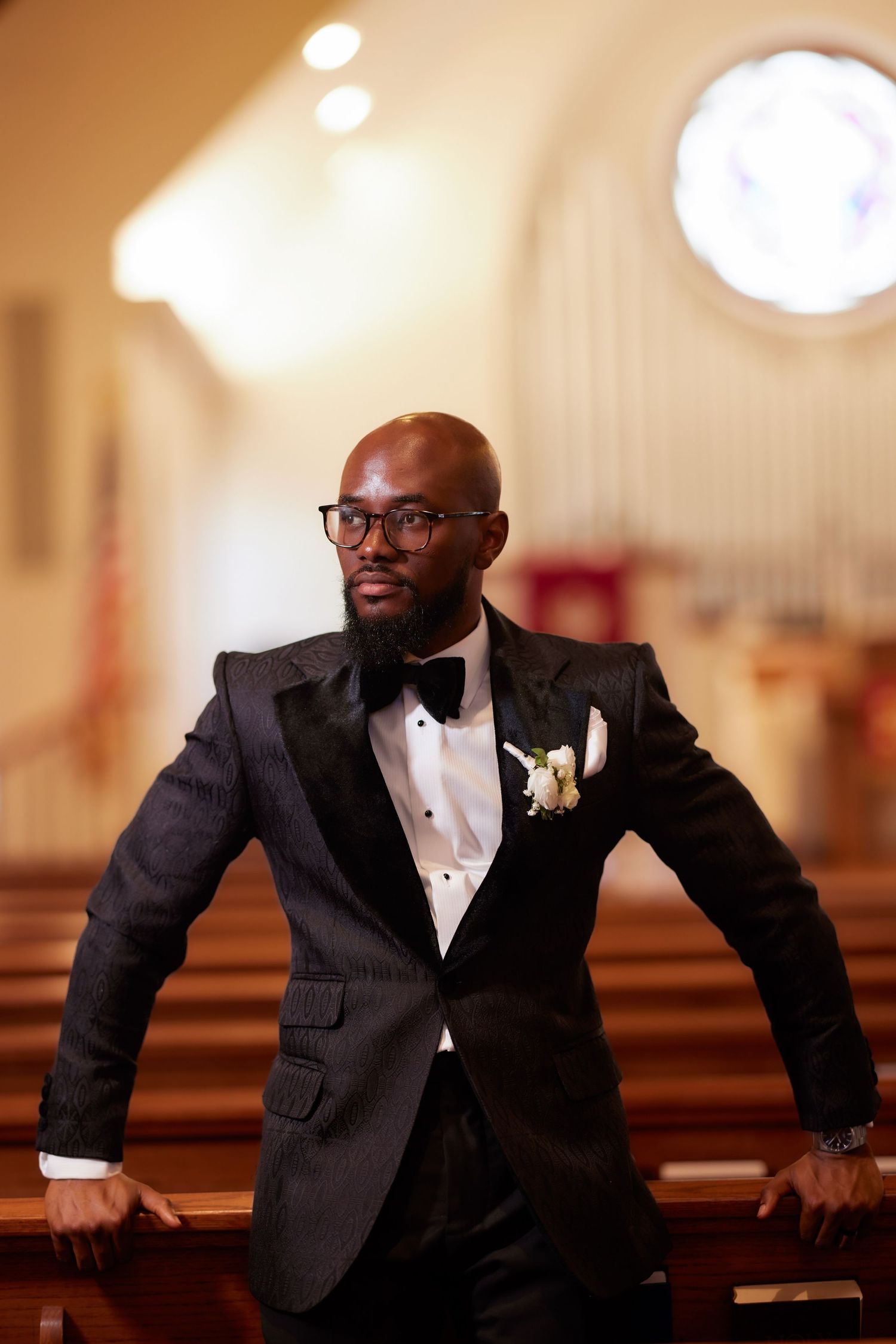 A groom in a black tuxedo and glasses sits in a church pew while smiling warmly and appearing joyful.
