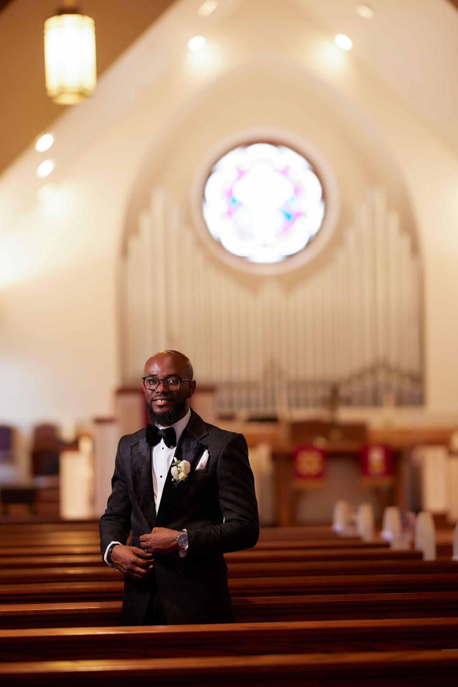 A person in a black tuxedo stands smiling in a church sanctuary with wooden pews and a round stained glass window.