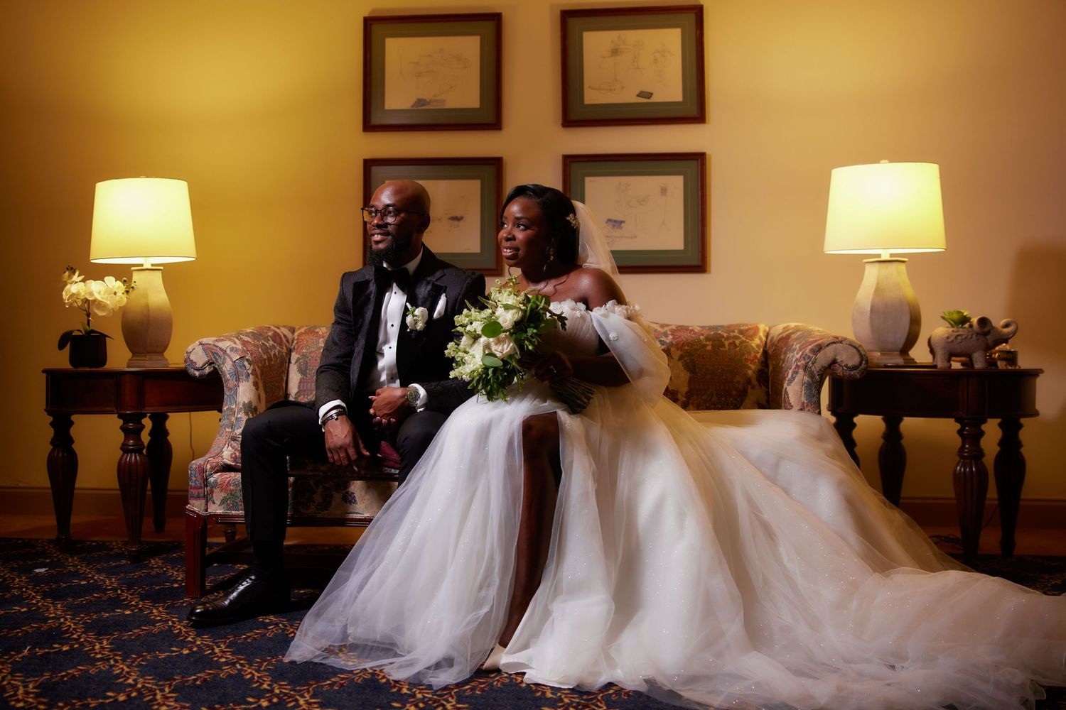 Newlyweds in flowing white dresses walk side by side through a golden-lit hallway at their wedding reception.