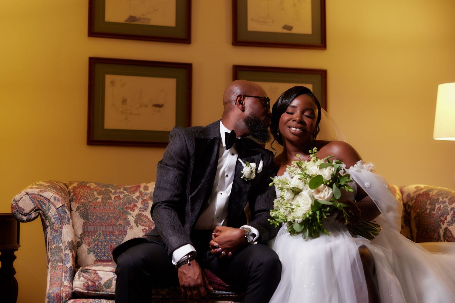 Wedding guests share an intimate moment during a reception in a warmly lit room with framed pictures on the wall.